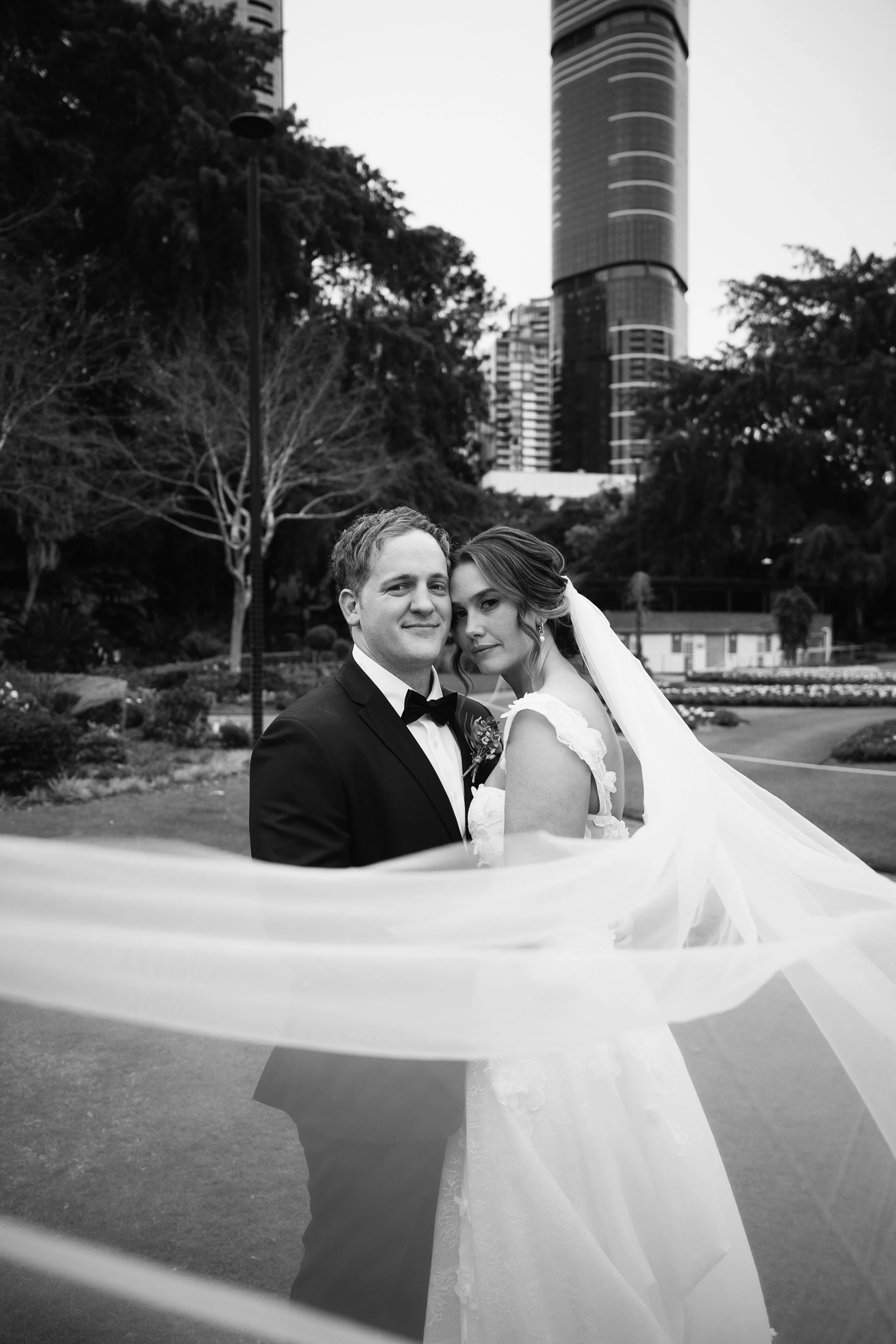 Black and white photo of a bride and groom standing together outdoors in a city park, with tall buildings in the background. The groom wears a tuxedo and the bride wears a wedding gown with a veil.