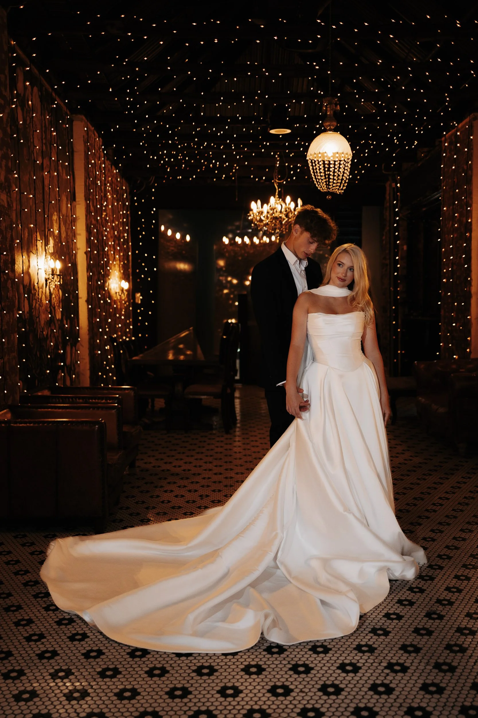A bride and groom standing together in a dimly lit, decorated venue with fairy lights and chandeliers, the bride in a white wedding gown with a long train and the groom in a black suit with a white shirt.