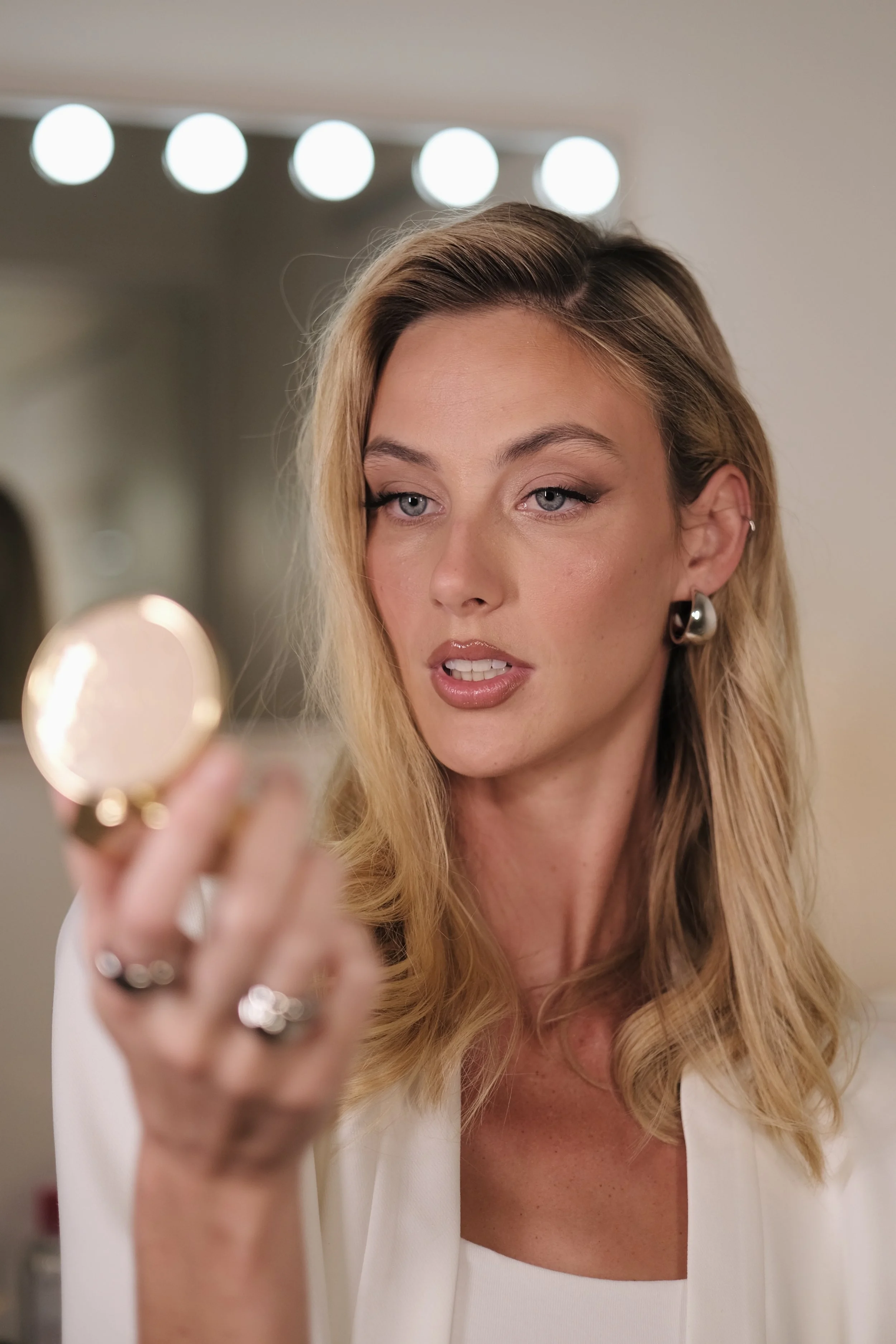 A woman with blonde hair and earrings looking into a makeup compact mirror.