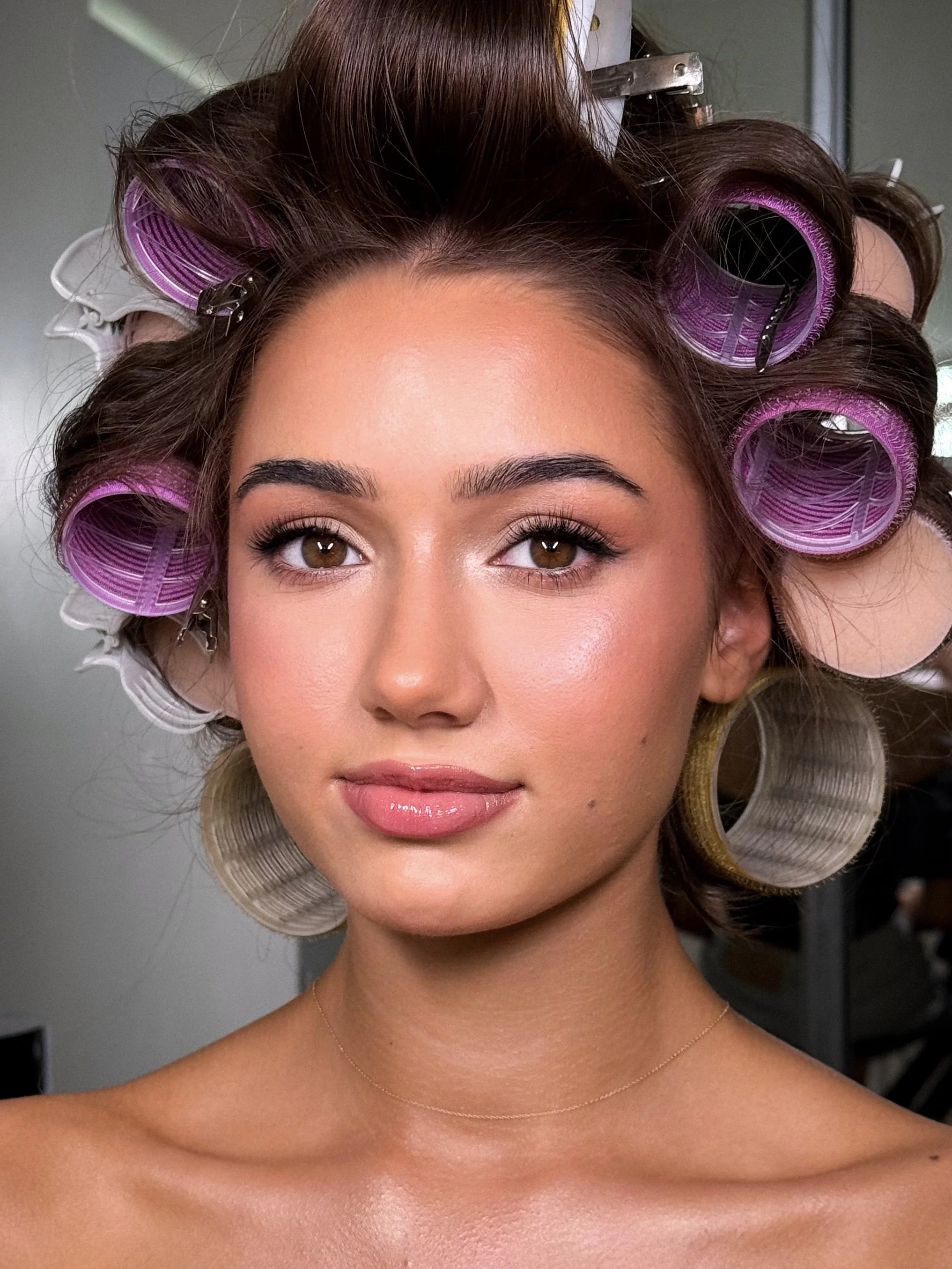 A young woman with brown hair in curlers looks directly at the camera with a neutral expression.