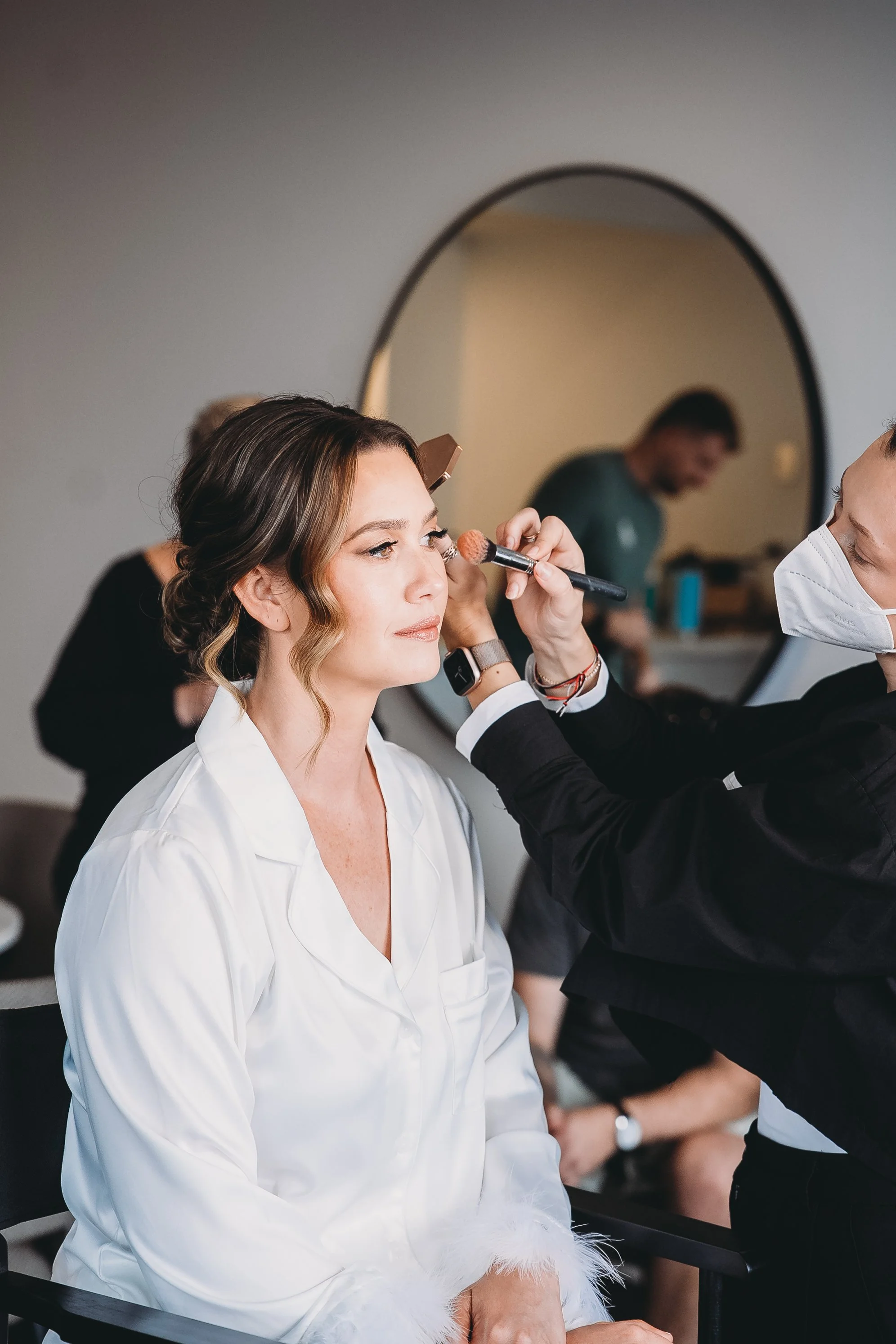 A woman with styled hair and a white silk robe sits while a makeup artist applies makeup on her face, reflected in a round mirror.
