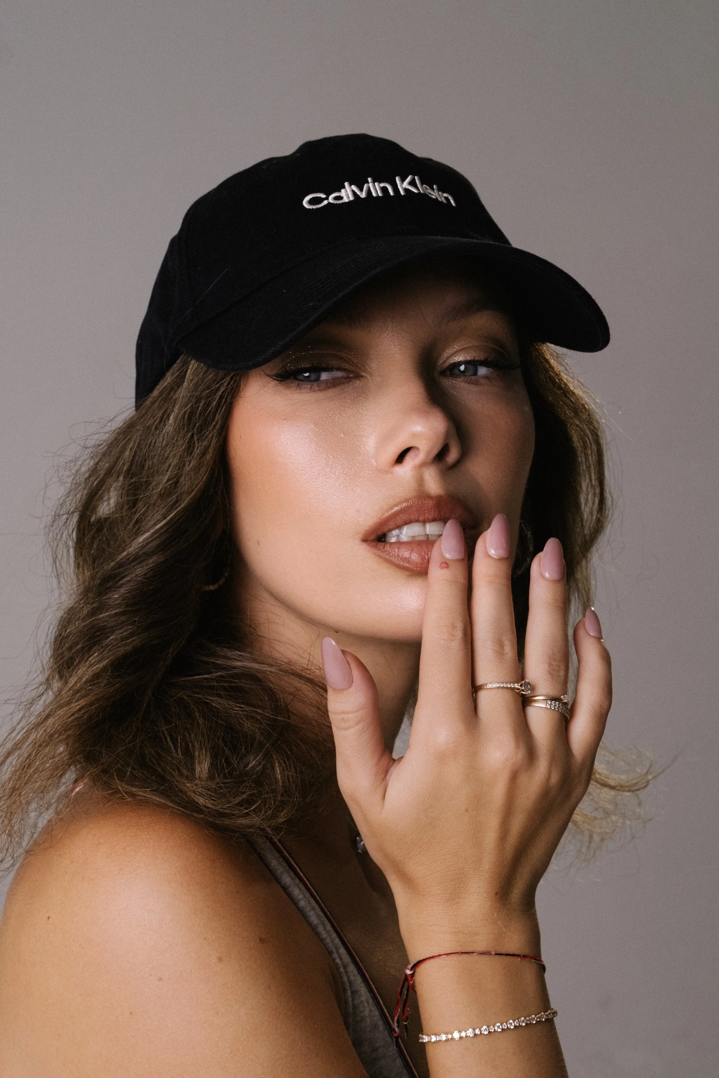 A woman wearing a black Calvin Klein baseball cap, with wavy brown hair, slightly parted lips, and intricately manicured nails, with her fingers near her lips.