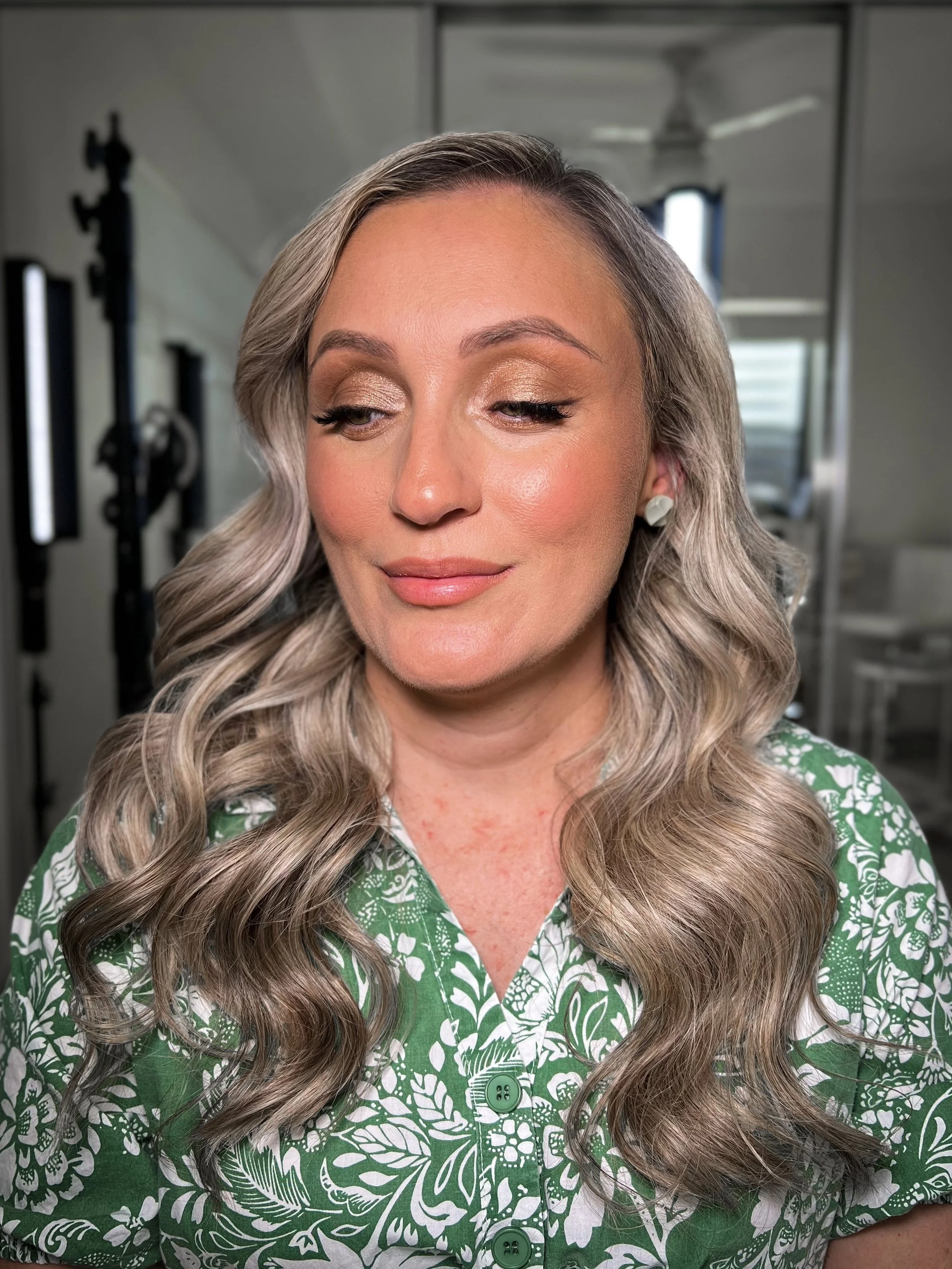 Close-up of a woman with styled wavy blonde hair, wearing makeup, and a green patterned blouse, indoors in a room with professional equipment and a mirror in the background.
