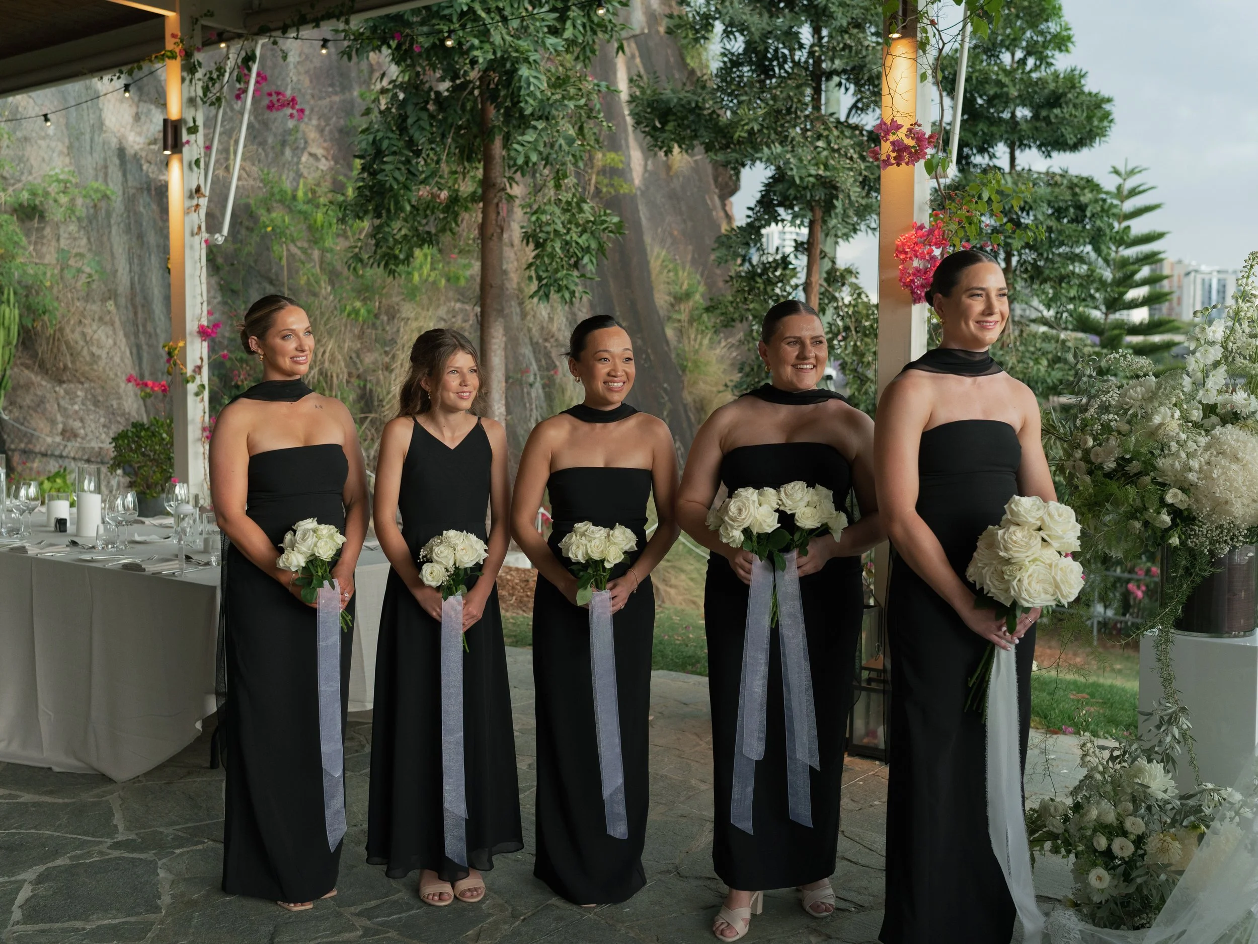 Five women in black evening dresses holding white roses, standing side by side at an outdoor event with tables, plants, and flowers in the background.