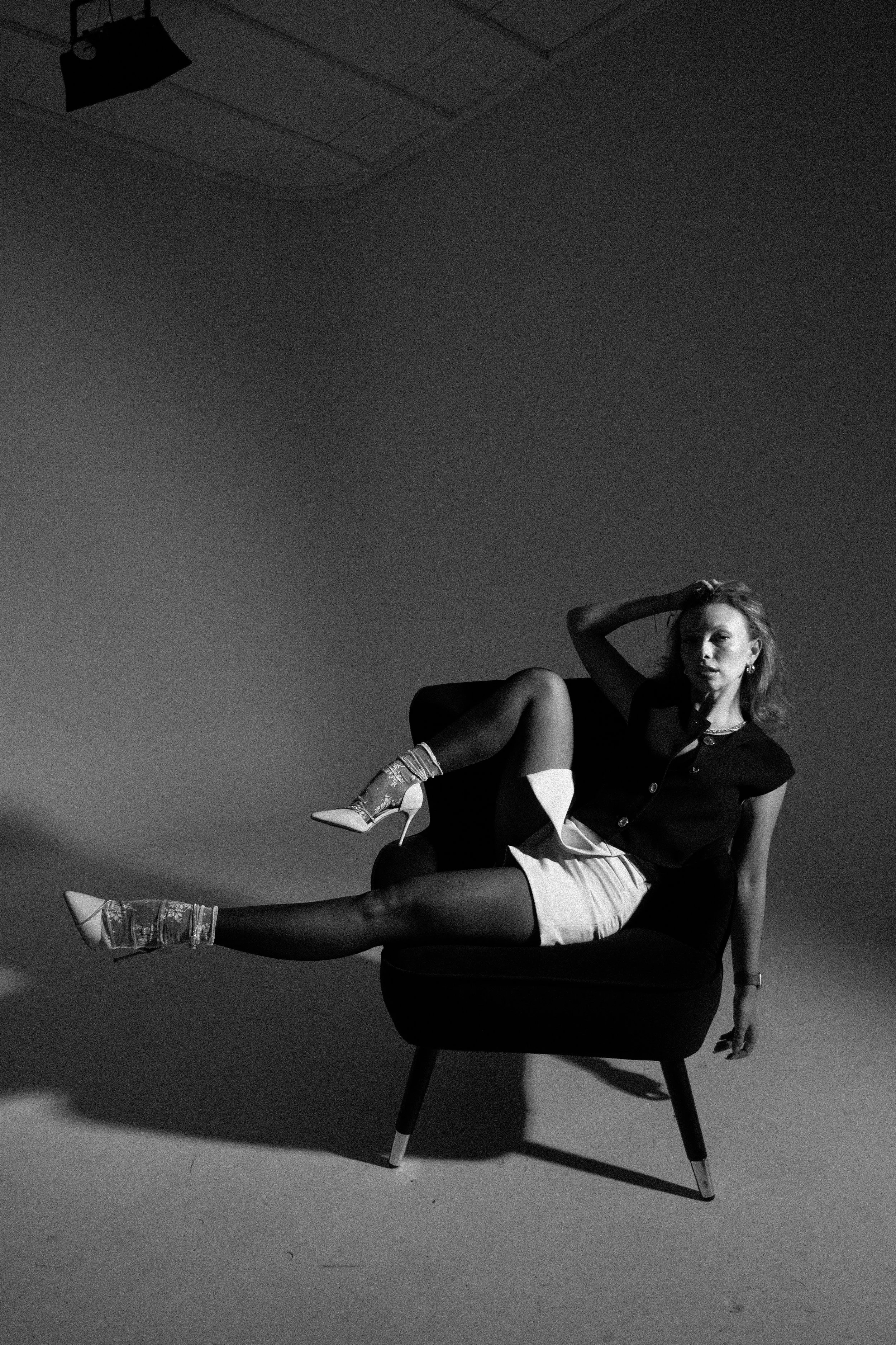 Black and white photo of a woman reclining on a chair with one leg extended and the other bent, wearing high heels, a dark top, and light-colored shorts, in a studio setting with a floor and ceiling visible.