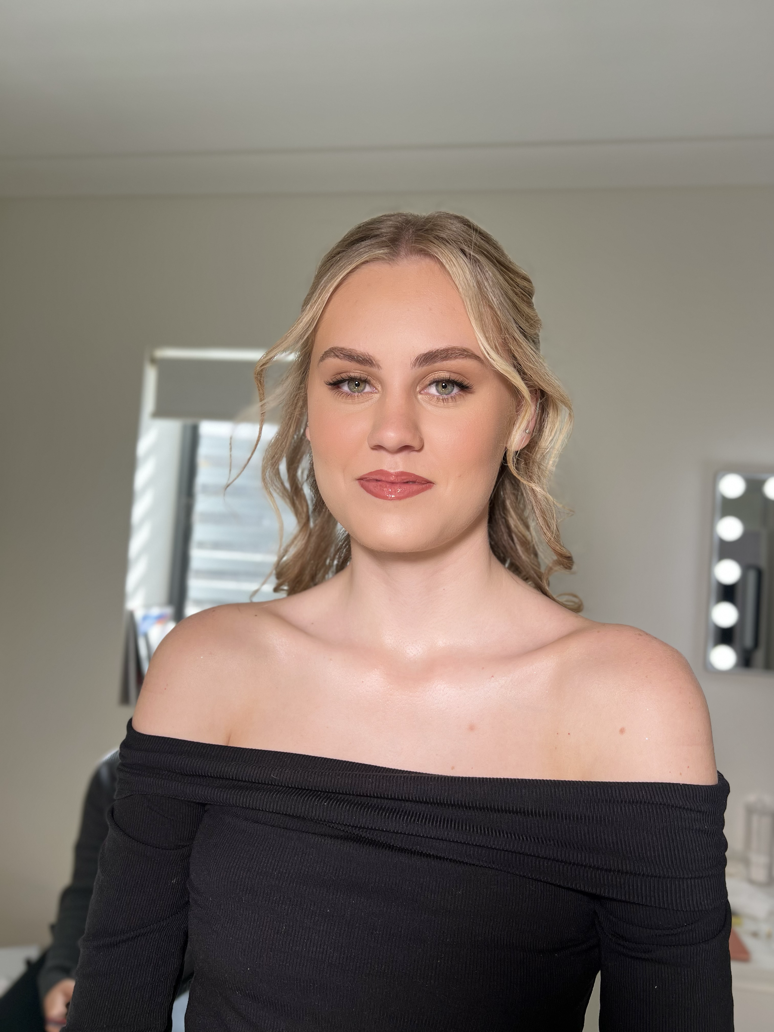 A young woman with blonde hair styled in loose waves, wearing an off-the-shoulder black top, smiling softly in a well-lit room with makeup mirrors and windows in the background.