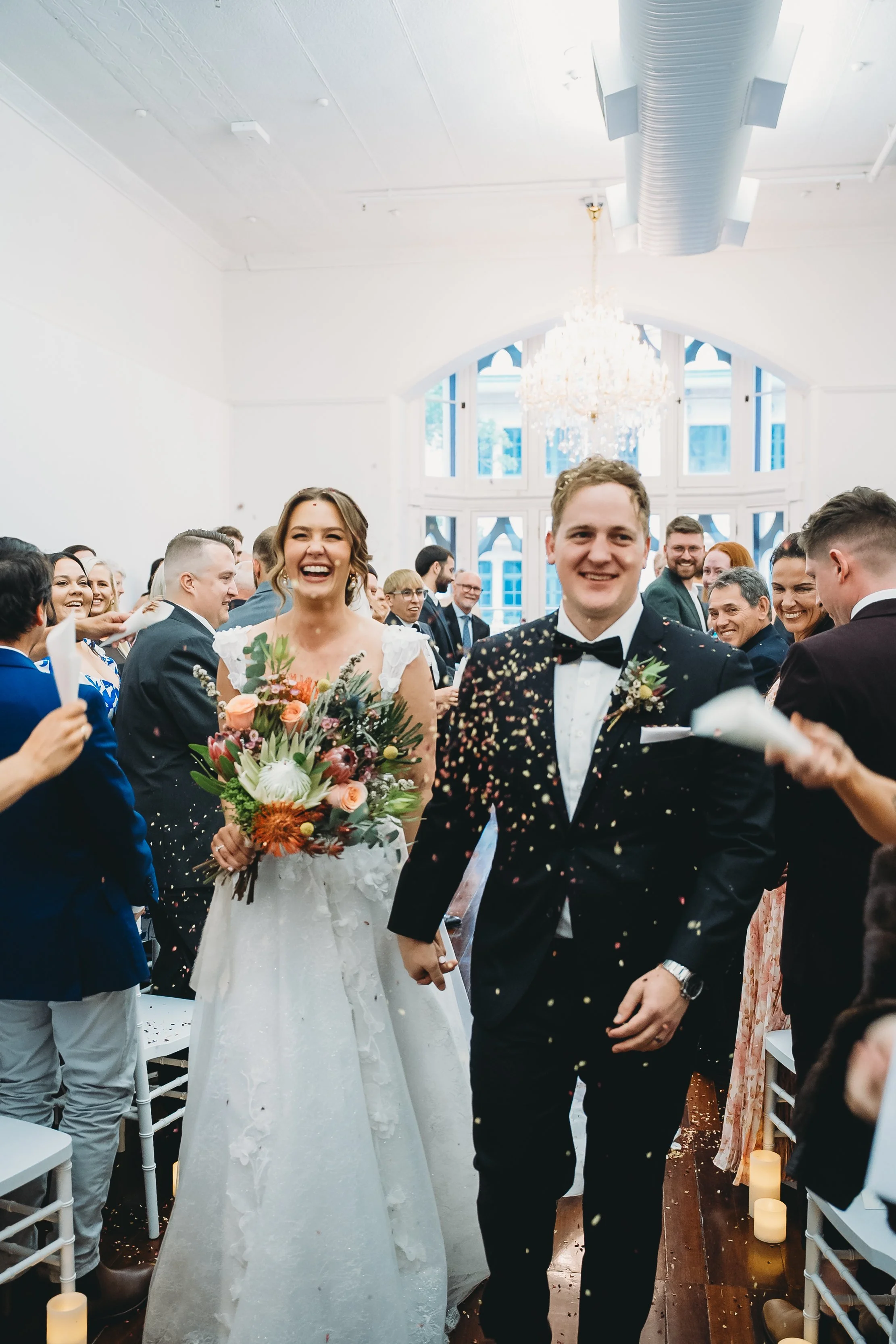 A newlywed couple walks down the aisle at their wedding reception, celebrating with guests throwing confetti. The bride is holding a colorful bouquet, and the groom is dressed in a tuxedo with a bow tie. The room is bright with large windows and a ch