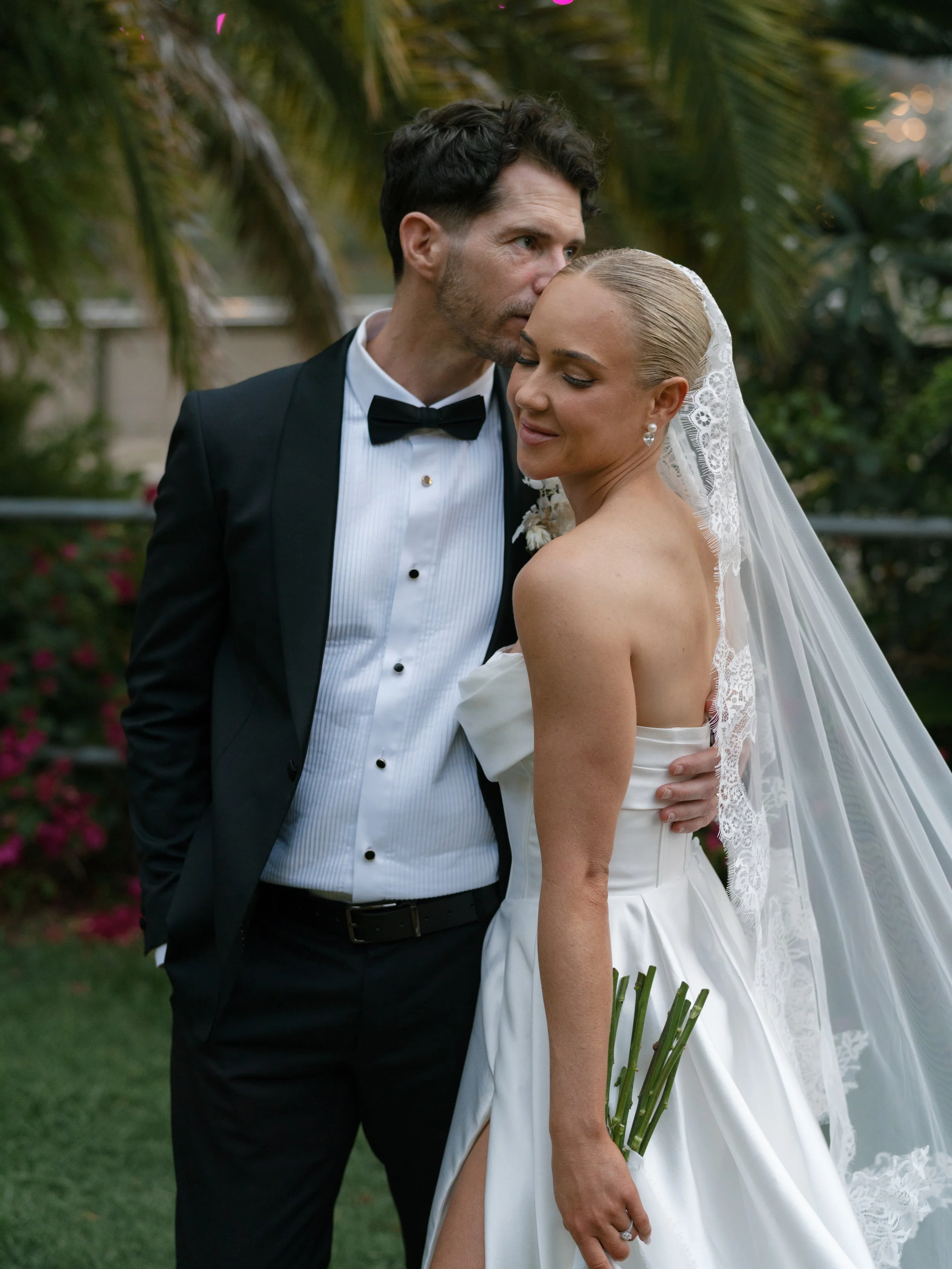 A bride and groom embrace outdoors on their wedding day. The groom, in a black tuxedo with a bow tie, leans in to kiss the bride's temple. The bride, in a strapless white wedding gown with a veil, holds a small bouquet of green stems and has blonde h