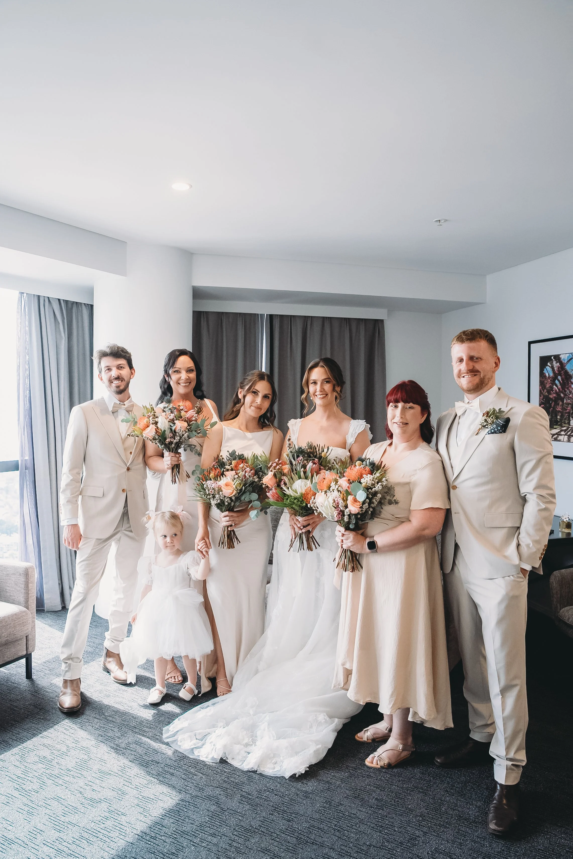 A wedding party of six adults and one young girl, dressed in formal attire, posing indoors. The women are holding bouquets of flowers, and the men are in suits with ties. A bride in a white wedding gown is in the center. The setting appears to be a m