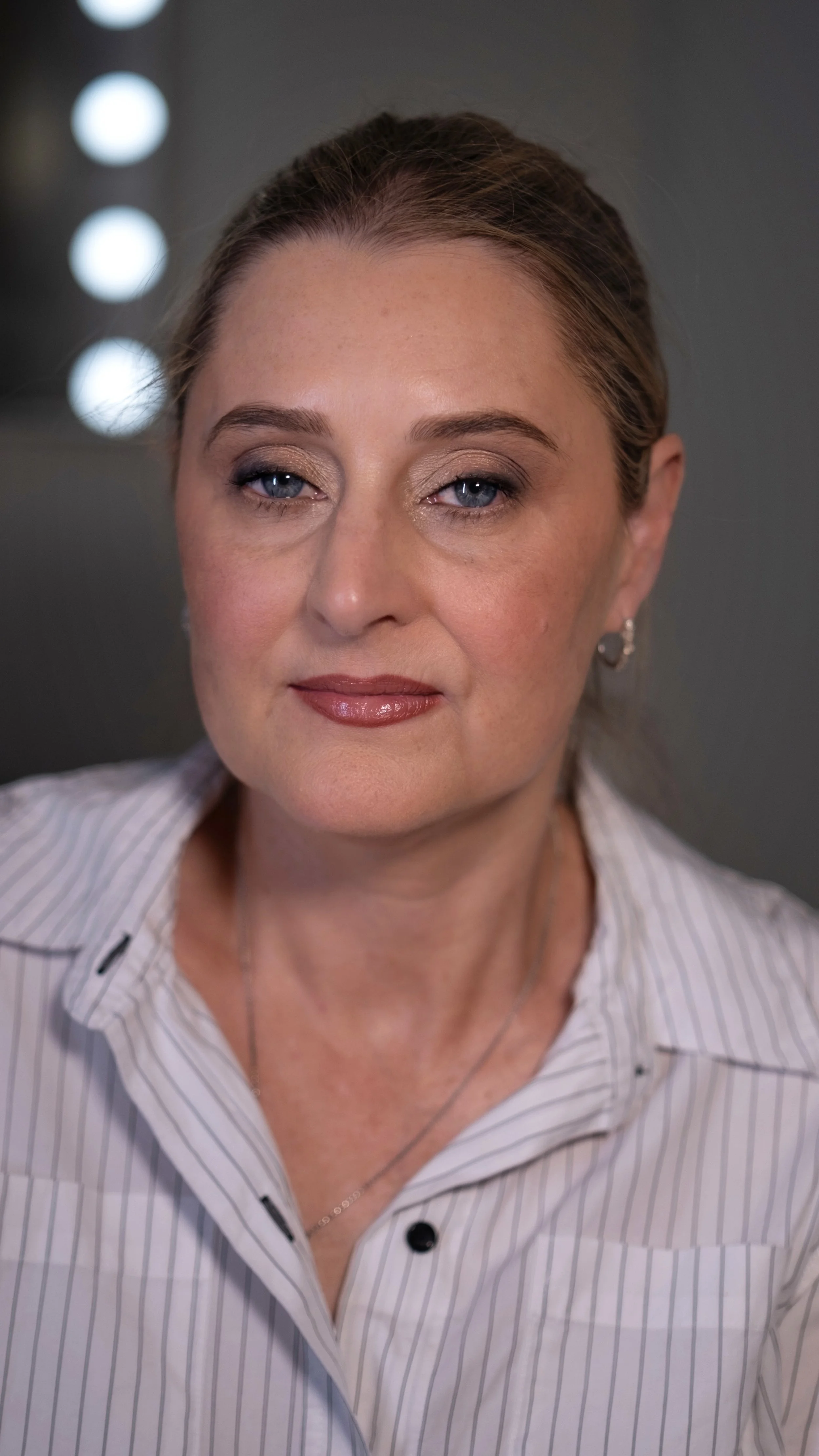 A woman with blonde hair, blue eyes, wearing a white striped shirt, earrings, and a necklace, is looking directly at the camera with a slight smile.