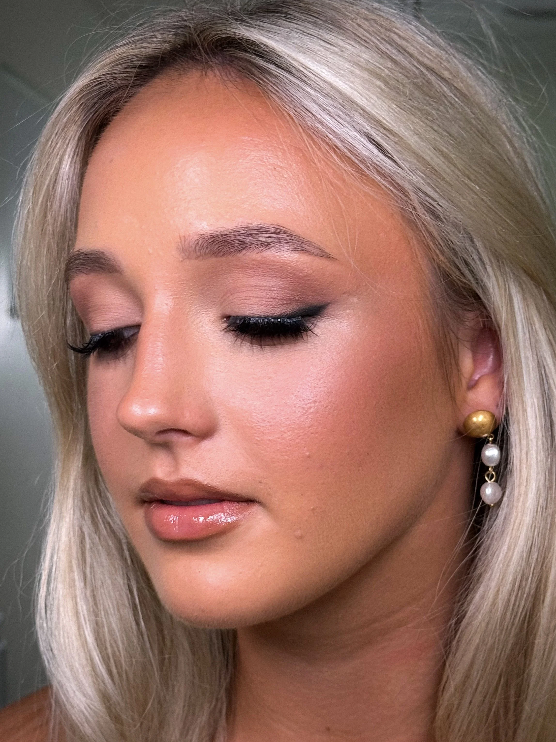 Close-up of a young woman with blonde hair, wearing makeup with pinkish eyeshadow, black eyeliner, and glossy lips, and gold and pearl earrings.