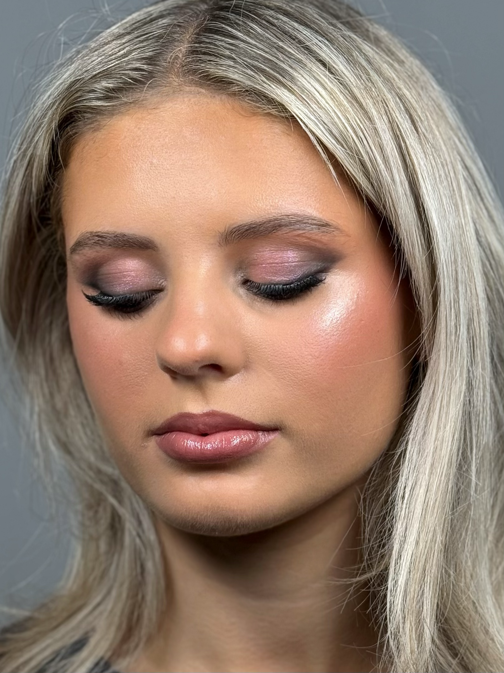 Close-up of a woman with blonde hair and makeup with closed eyes, pink eyeshadow, black eyeliner, and glossy lips.