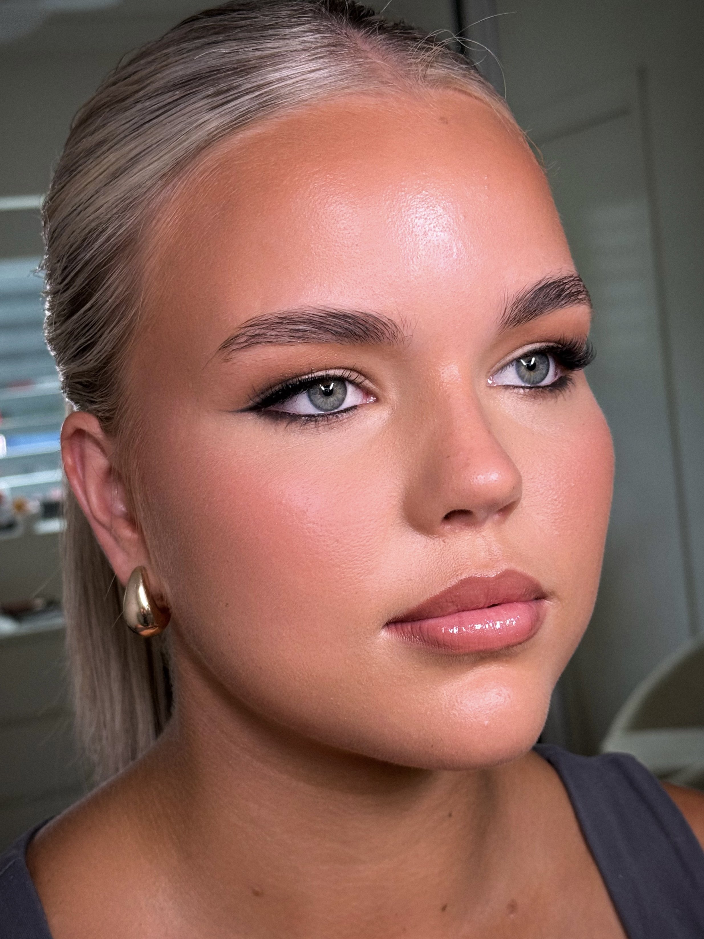A woman with blonde hair styled back, wearing gold hoop earrings, has flawless makeup with neutral eyeshadow, eyeliner, mascara, blush, and glossy lips. She is looking slightly to the side.