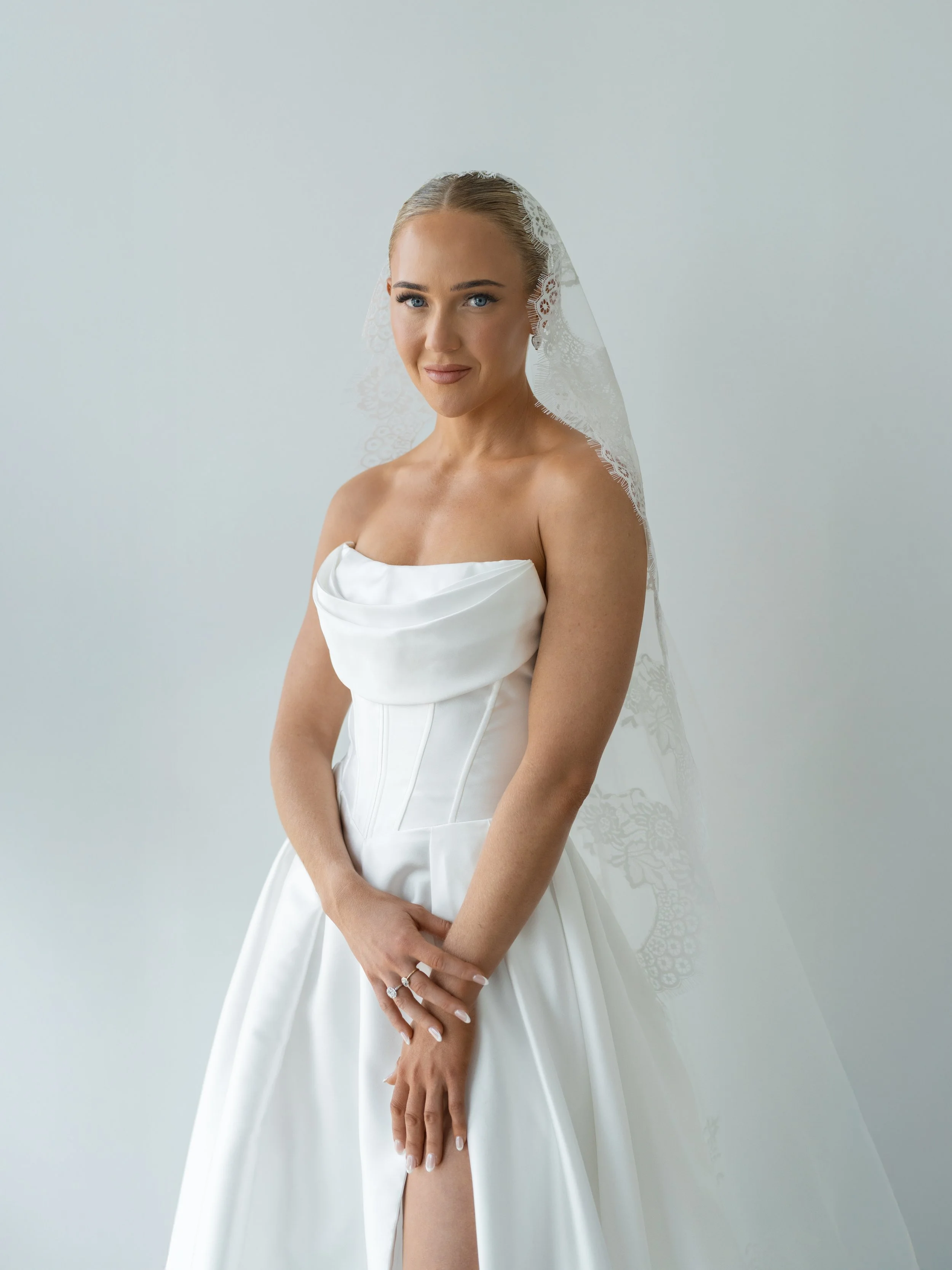 Bride in a white wedding gown with a lace veil, standing against a plain light background.