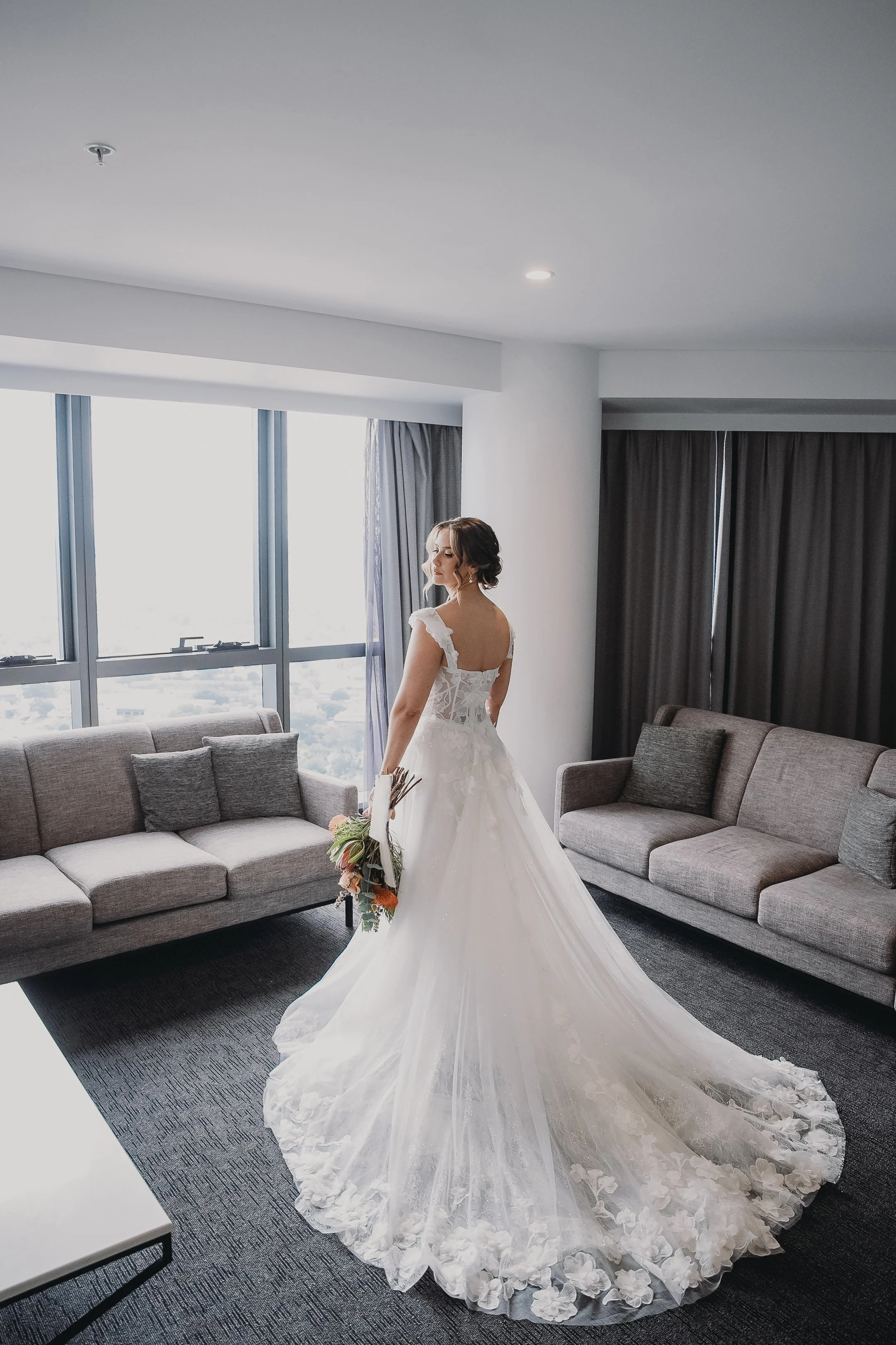 A bride in a white wedding dress with a long train, holding a bouquet, standing in a modern hotel room near large windows with gray couches and dark curtains.
