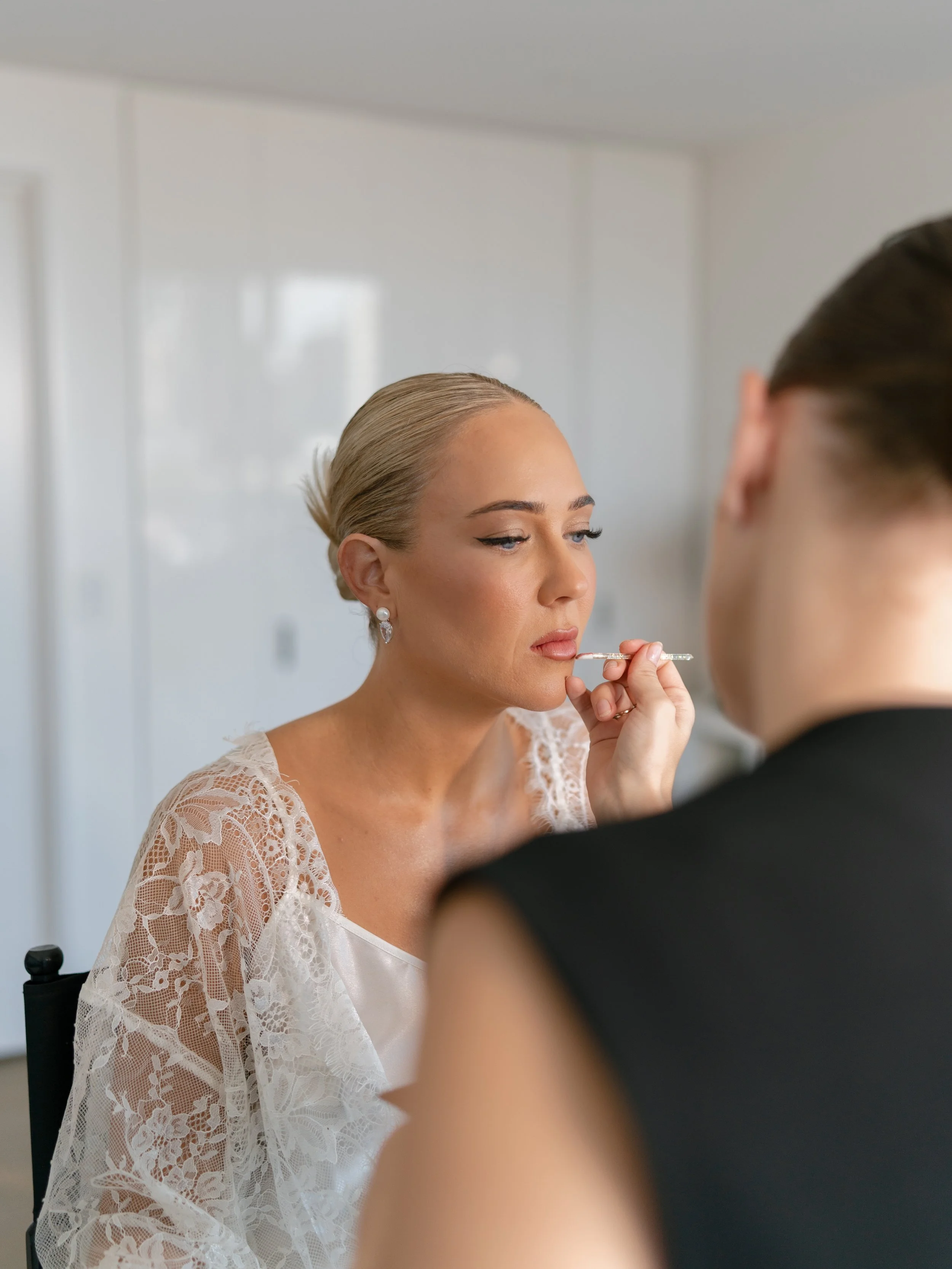 A woman with blonde hair in an updo, wearing pearl earrings and a lace garment, is having makeup applied with a brush by another person with dark hair, in a bright, clean room.