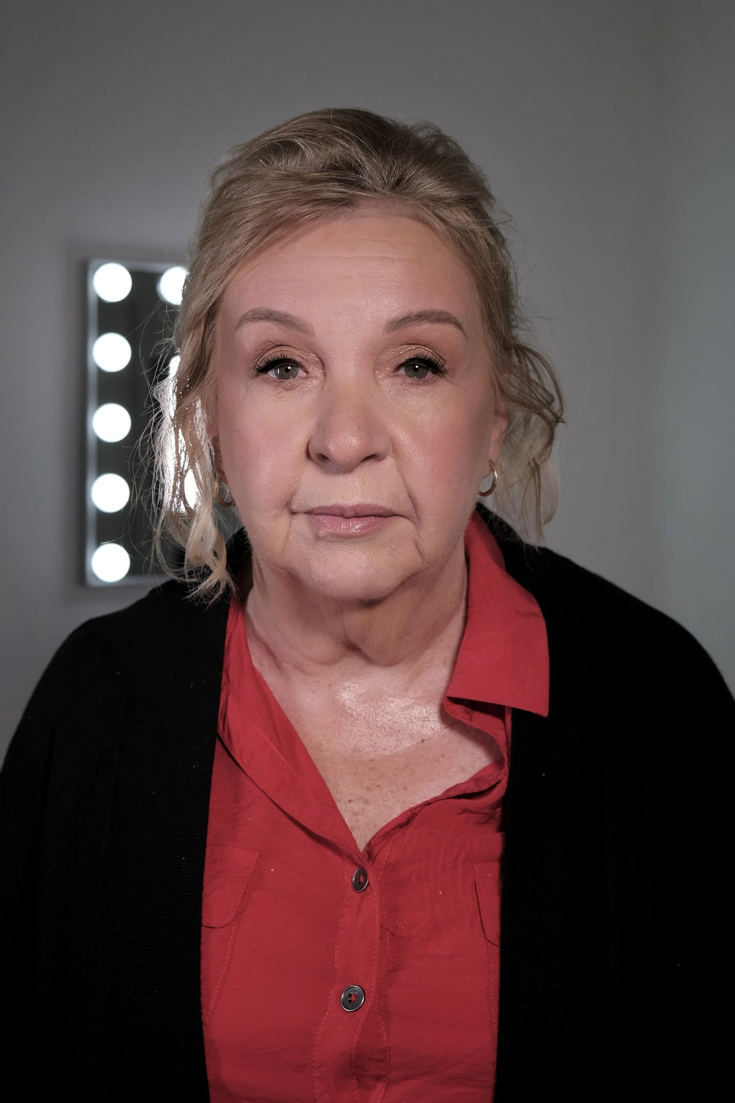A mature woman with blonde hair, wearing a red blouse and black sweater, looking directly at the camera with a neutral expression. A mirror with bright lights is visible in the background.