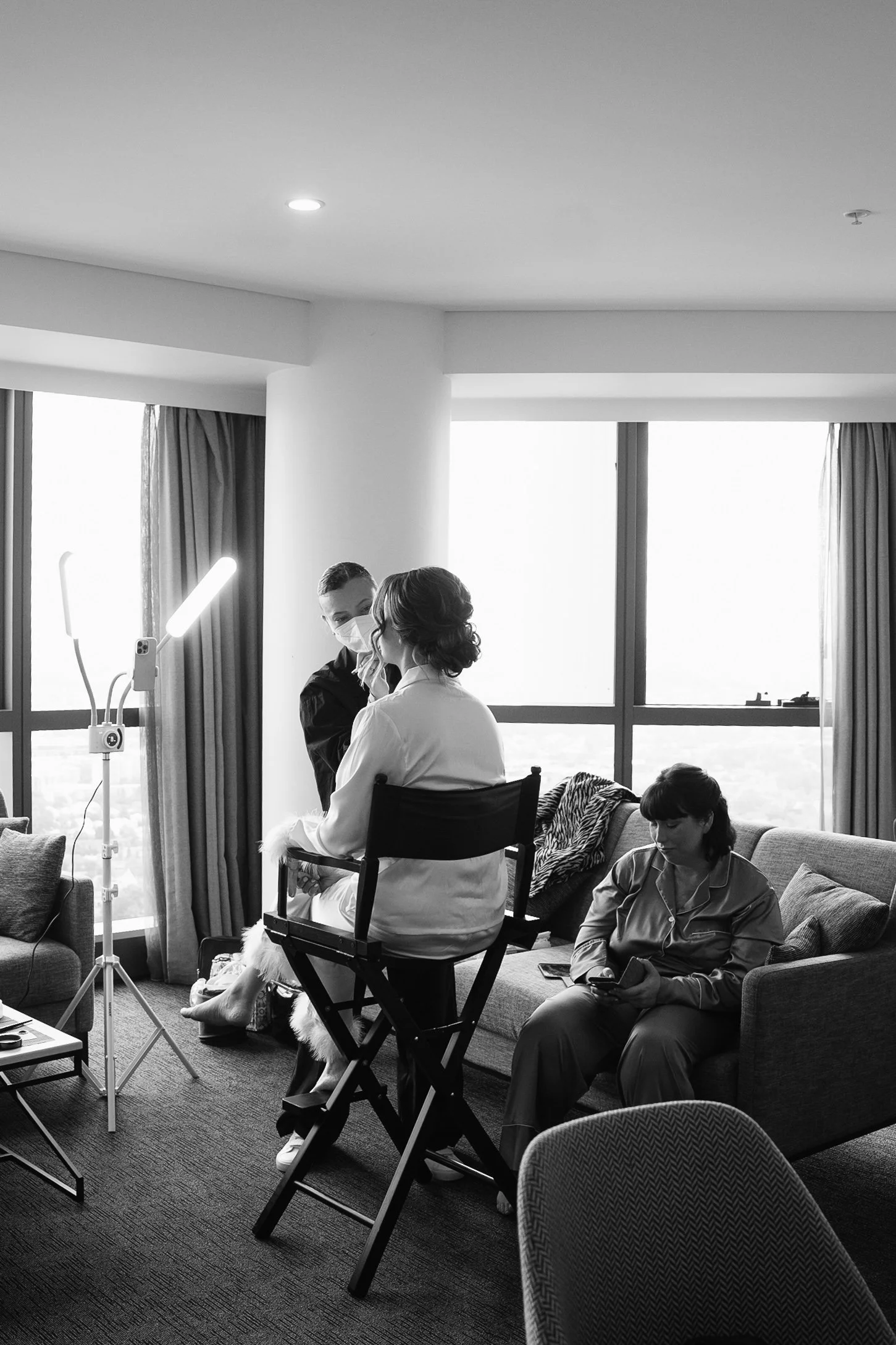 Three women in a hotel room, two sitting on a sofa and one on a director's chair, during a makeup or styling session, with large windows in the background.