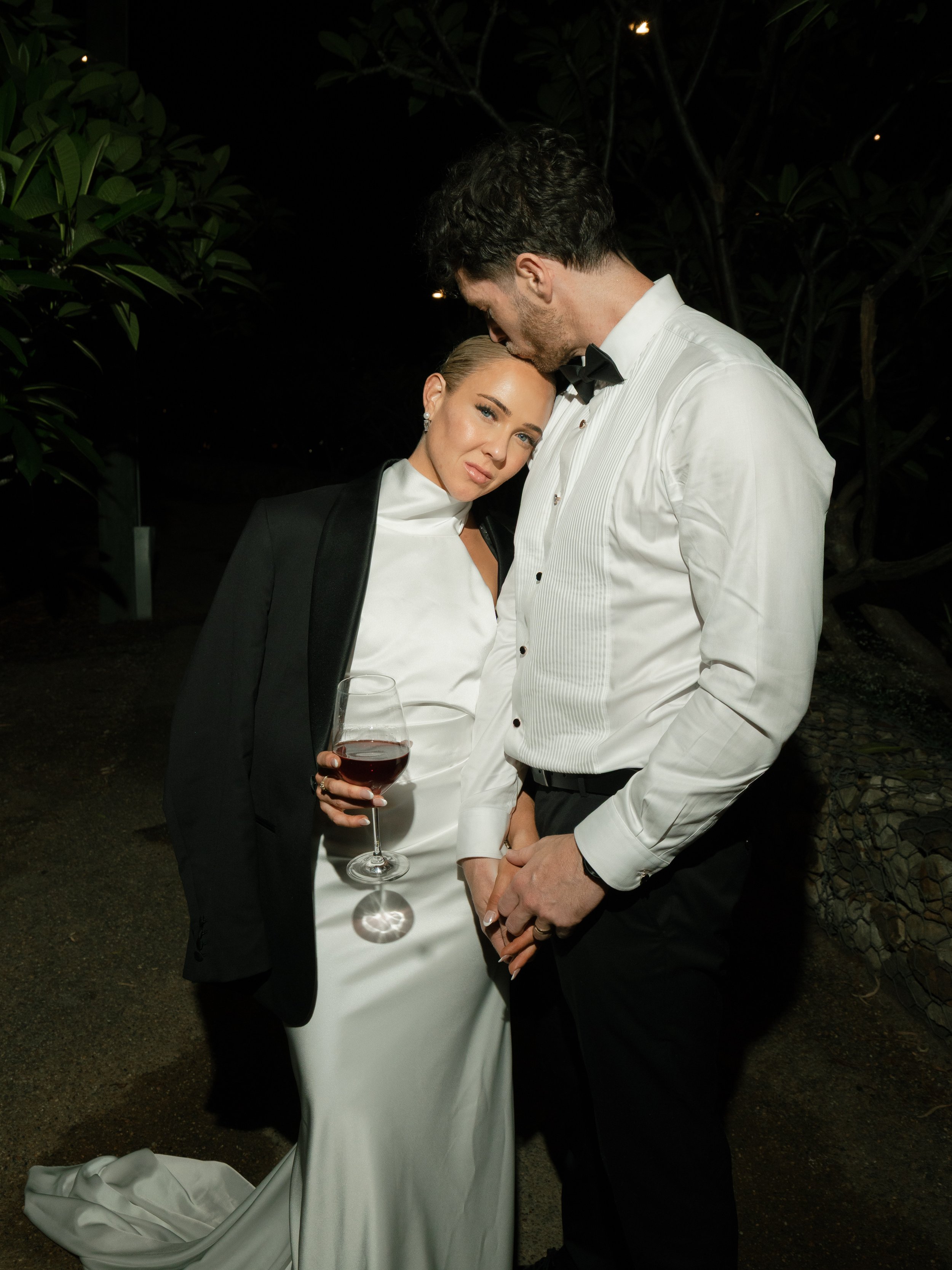 A couple dressed in formal attire, with the woman holding a glass of red wine, standing close together outdoors at night.