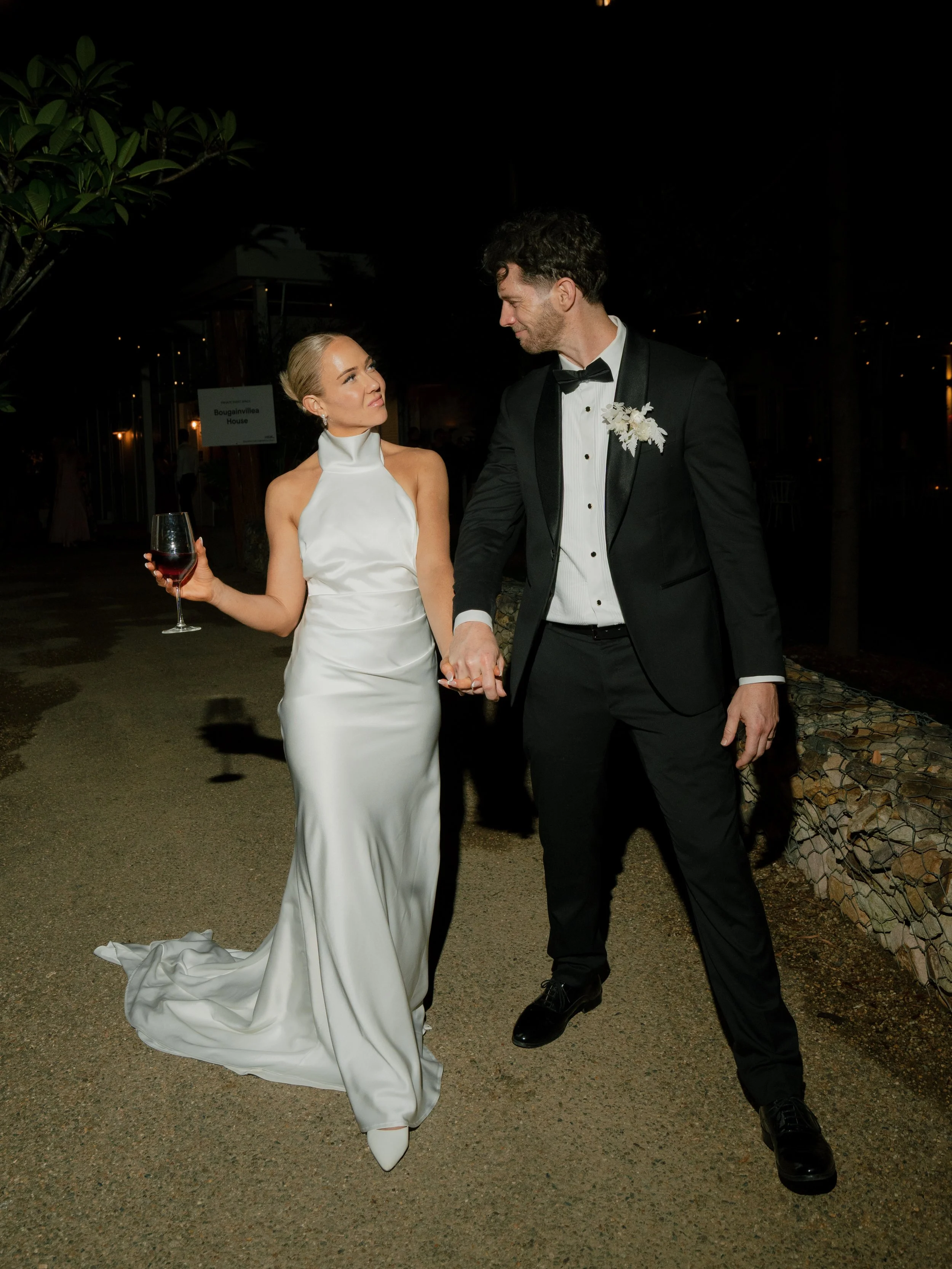 A bride and groom holding hands at night, dressed in formal wedding attire, with the bride holding a glass of red wine.