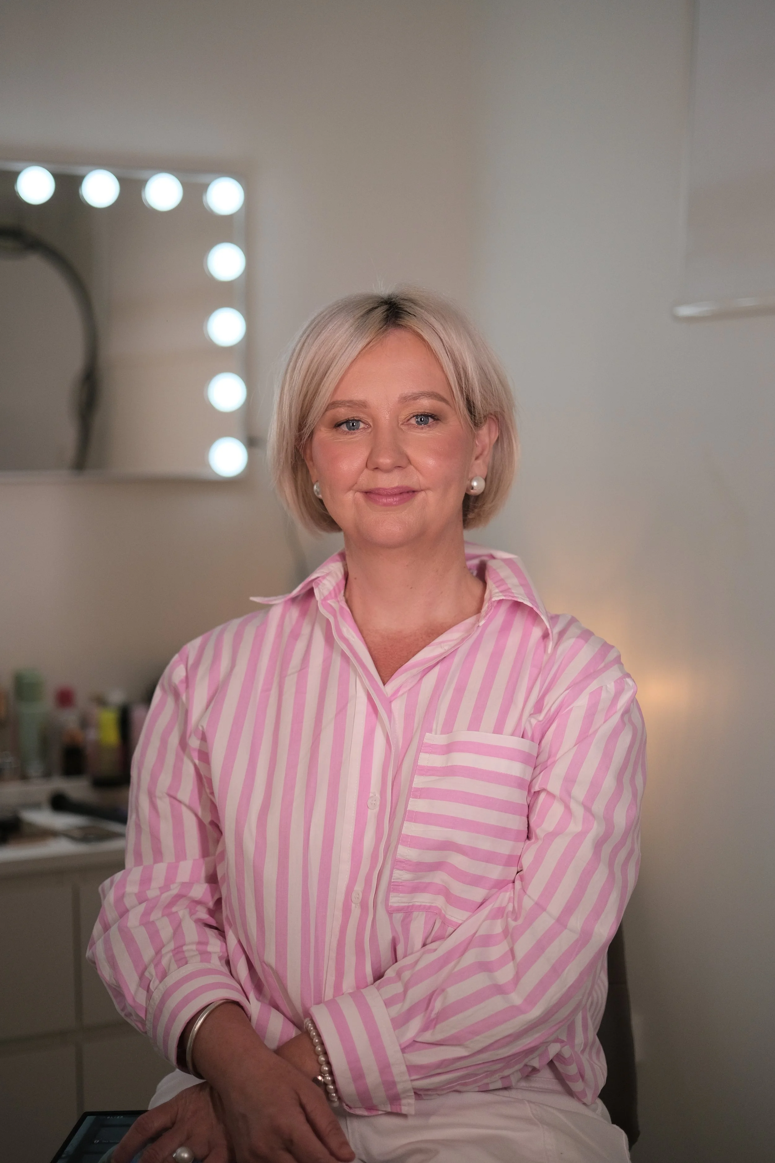 A woman with short blonde hair, wearing a pink and white striped shirt and pearl earrings, sitting in a room with a mirror and bright lights in the background.