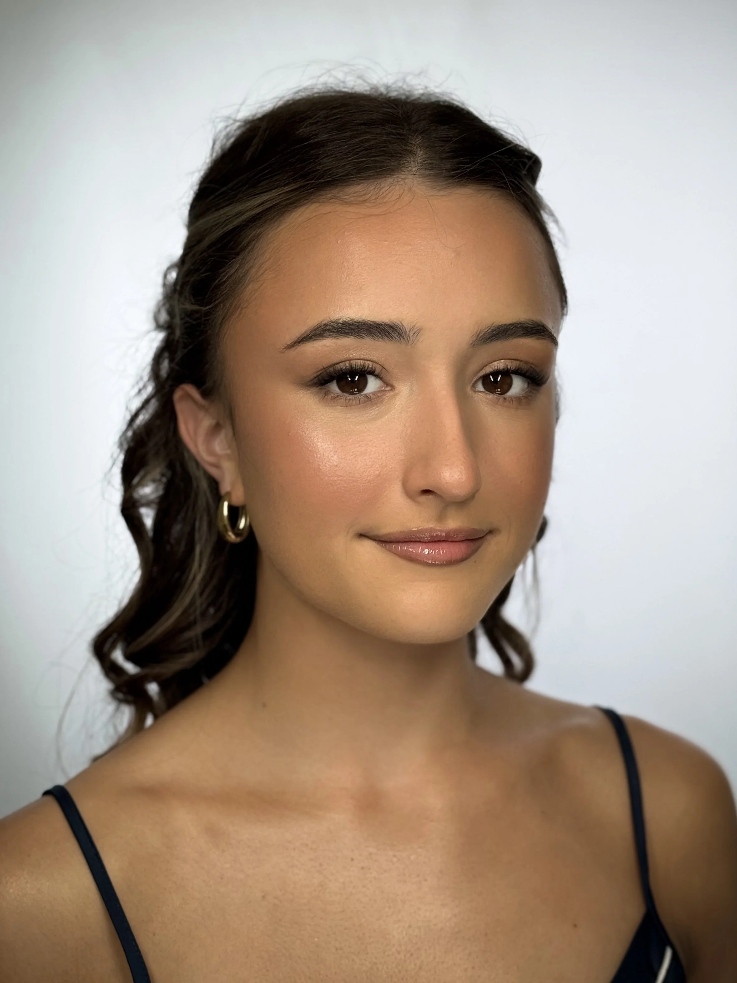 A young woman with medium brown, wavy hair, wearing hoop earrings and a dark spaghetti strap top, smiling softly against a plain white background.