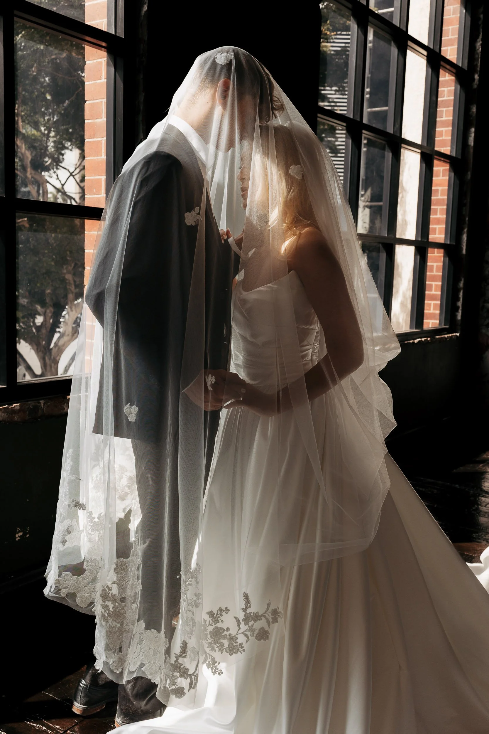 A bride and groom standing close together beneath a sheer veil, indoors near large windows with brick walls outside, sharing an intimate moment.