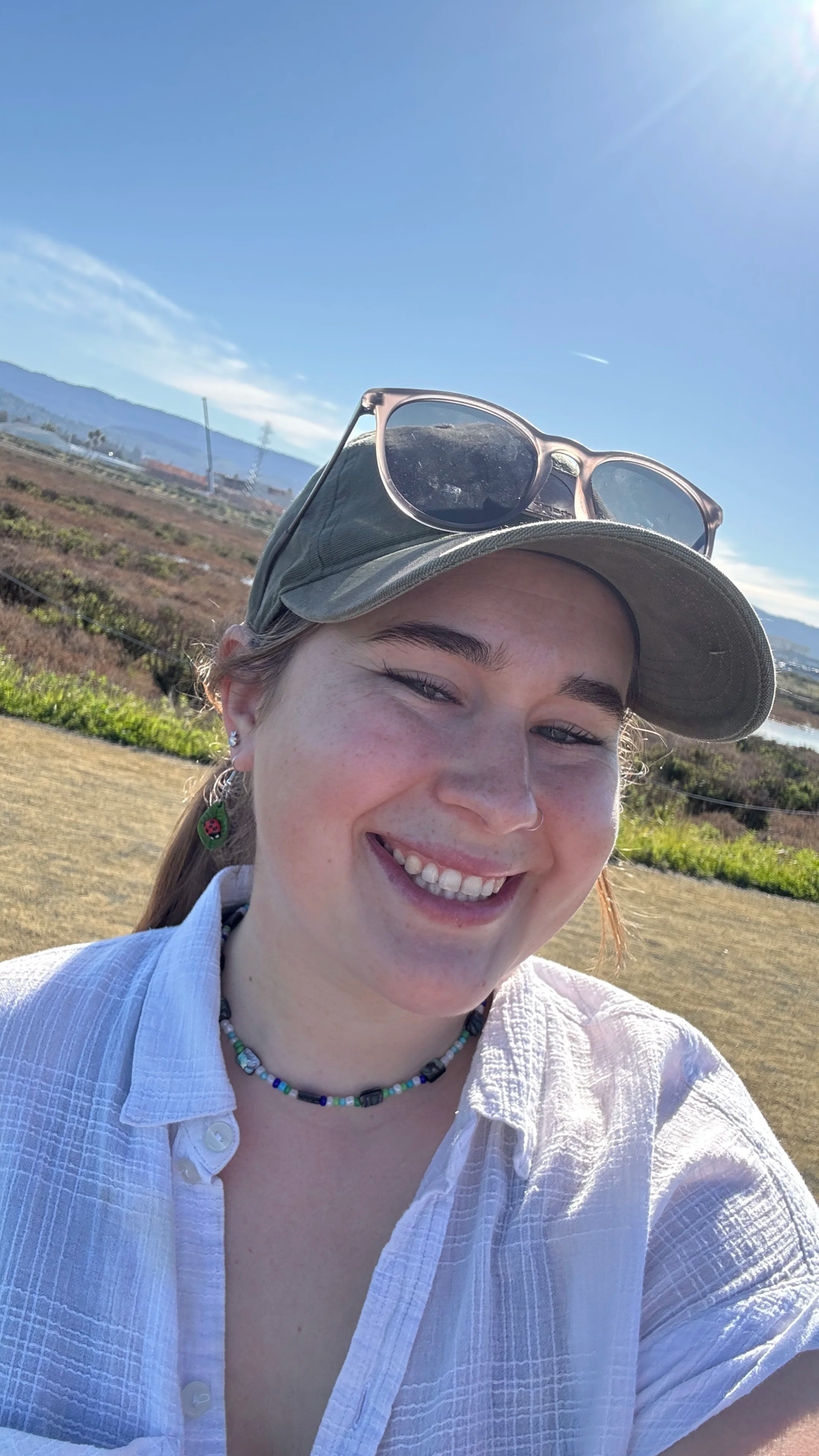 A woman smiling outdoors wearing a light-colored shirt, a green cap with sunglasses on top, and wearing colorful jewelry, with a landscape of fields, mountains, and a blue sky in the background.