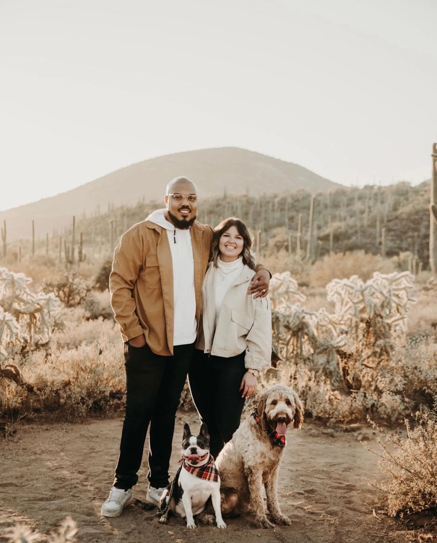 A man and woman standing together outdoors in a desert landscape with cacti and mountains in the background, along with two dogs. The man is wearing glasses, a tan jacket over a white hoodie, and black pants. The woman is dressed in a white jacket and black pants. Both are smiling and have their arms around each other.