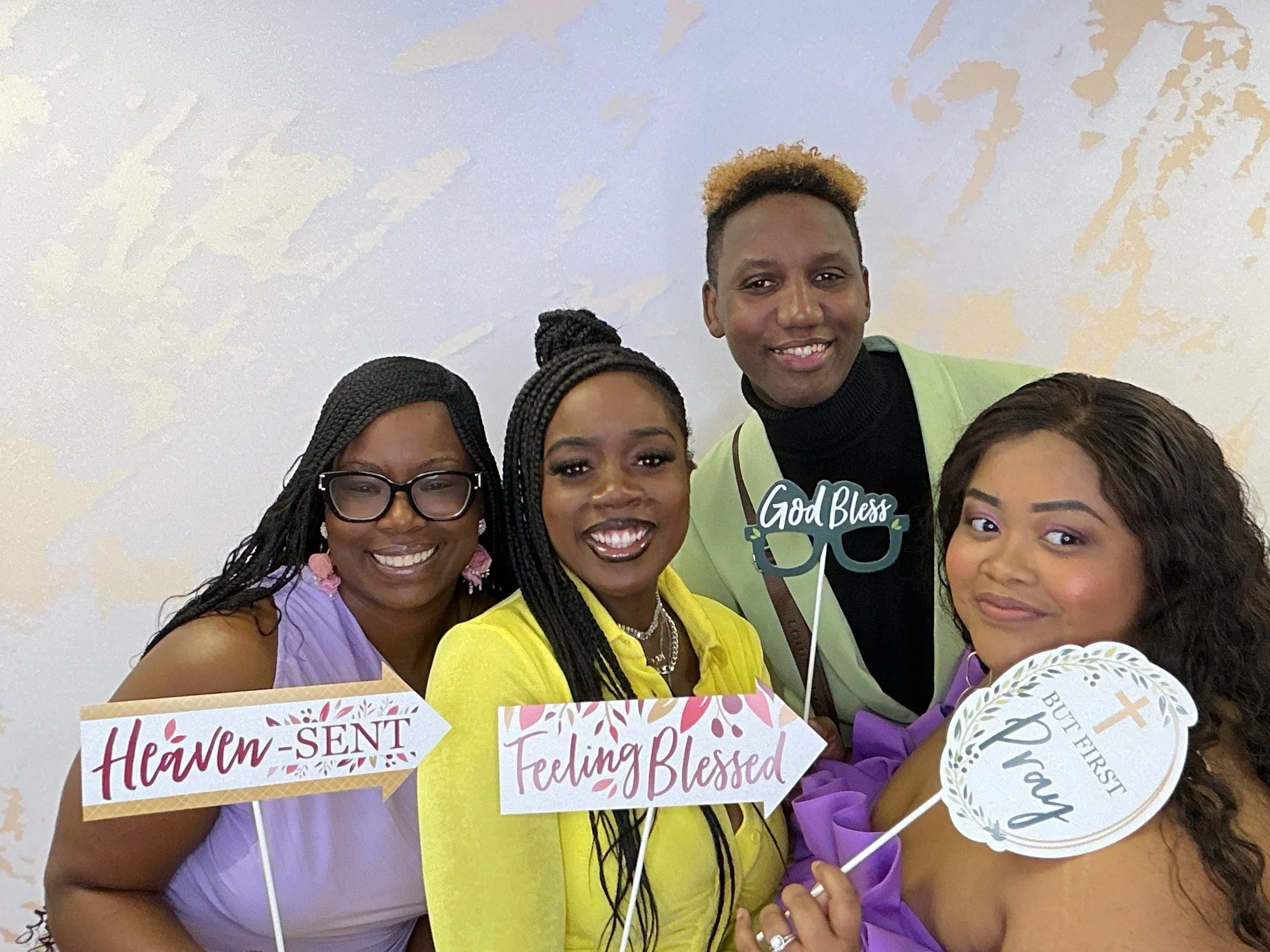 Four women and one man smiling at the camera at a celebration. They are holding signs with messages such as "Heaven-SENT," "Feeling Blessed," and "Pray," with the man also holding a prop that says "God Bless." The women wear purple, yellow, and light