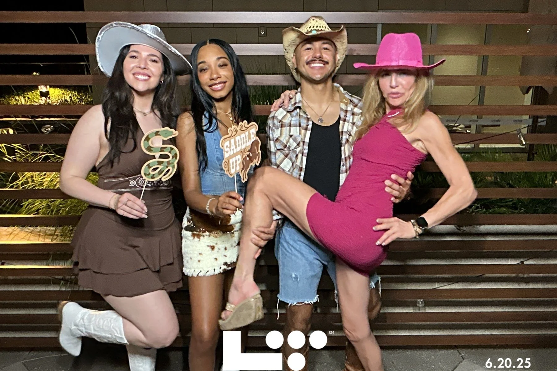 Group of four people dressed in Western and cowgirl costumes, posing outdoors at night with a wooden slatted background.