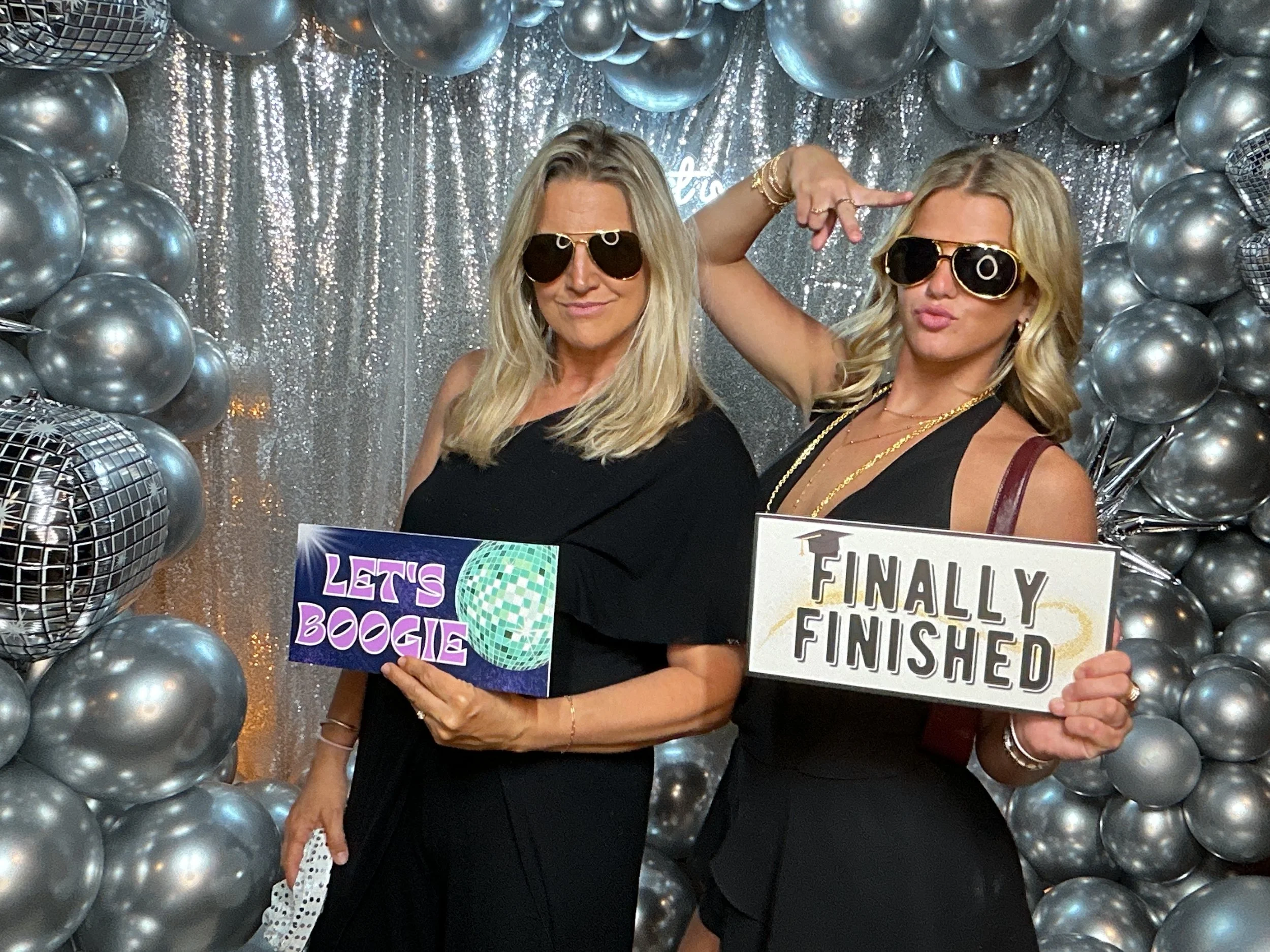 Two women with blonde hair wearing sunglasses and black dresses, posing with signs in front of a silver balloon backdrop at a celebration party. The woman on the left holds a sign that says 'Let's Boogie' with a disco ball illustration, and the woman