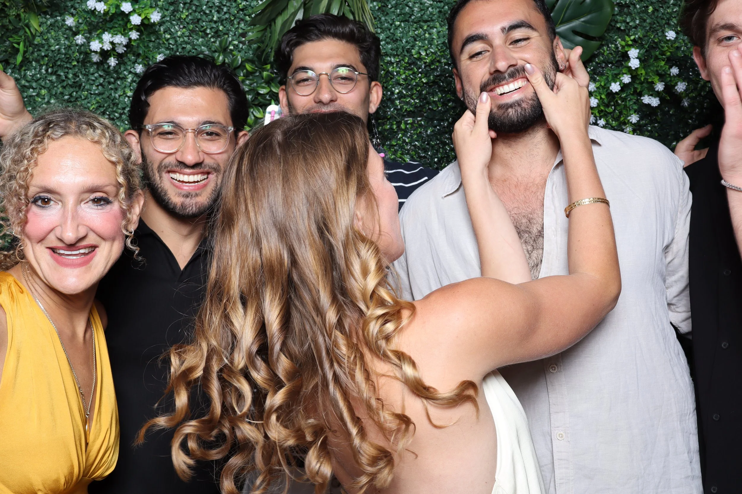 A group of people smiling and laughing, with a woman playfully covering a man's eyes, standing in front of a green leafy background.