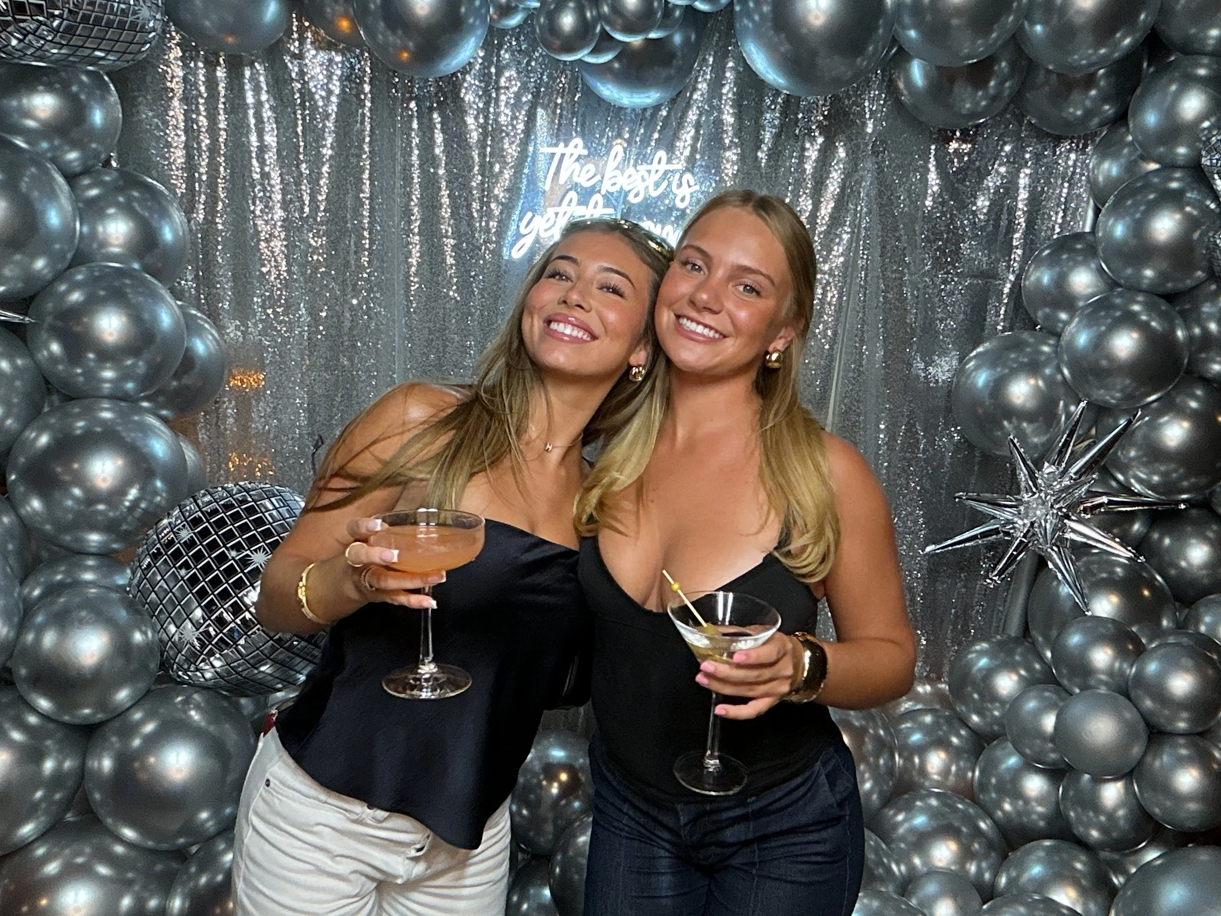 Two women celebrating at a party with silver balloons and a glittery backdrop, holding cocktails and smiling.