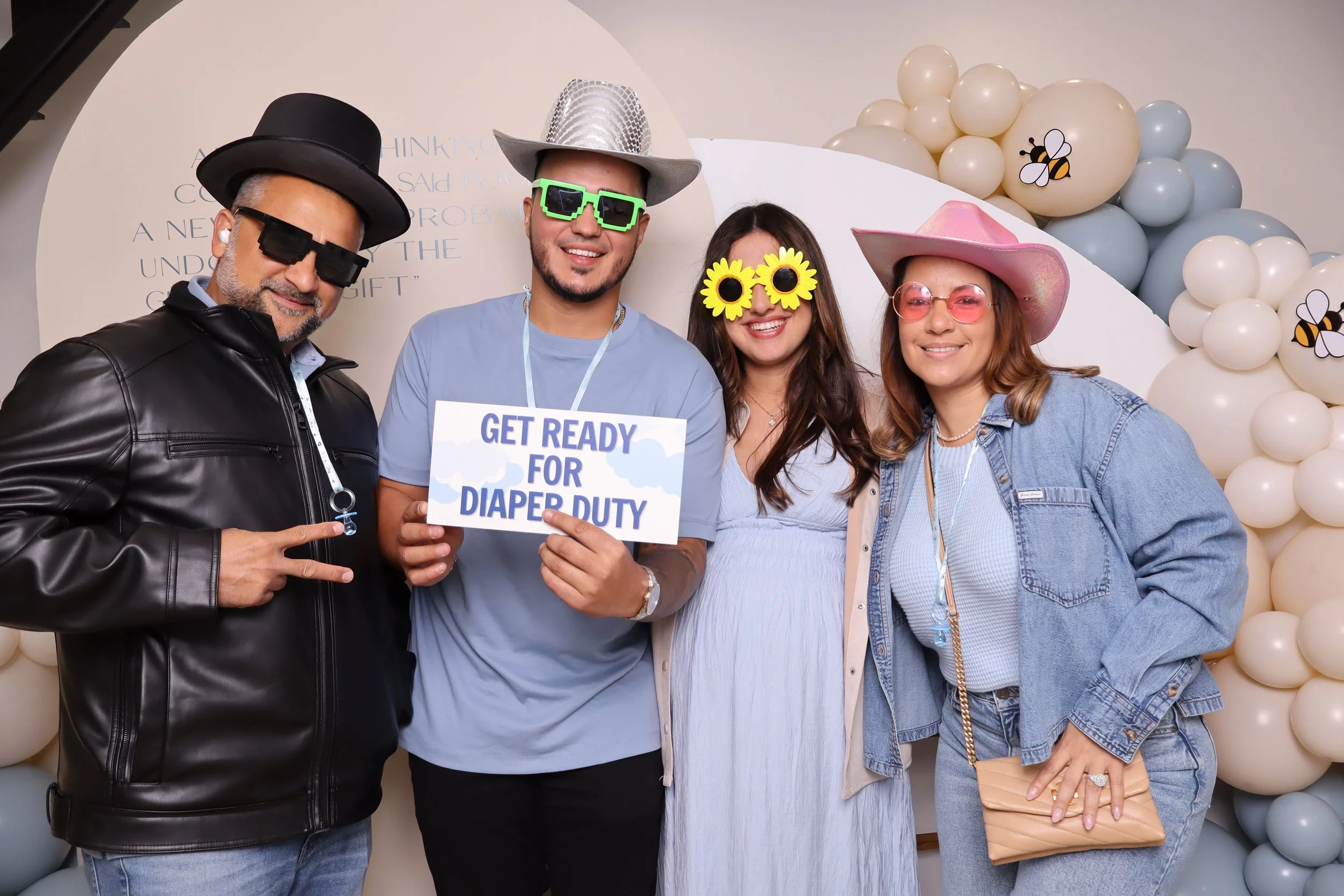 Four people dressed in playful attire with sunglasses and hats, holding a sign that reads 'Get Ready for Diaper Duty,' standing in front of a decorative backdrop with balloons and bee illustrations.