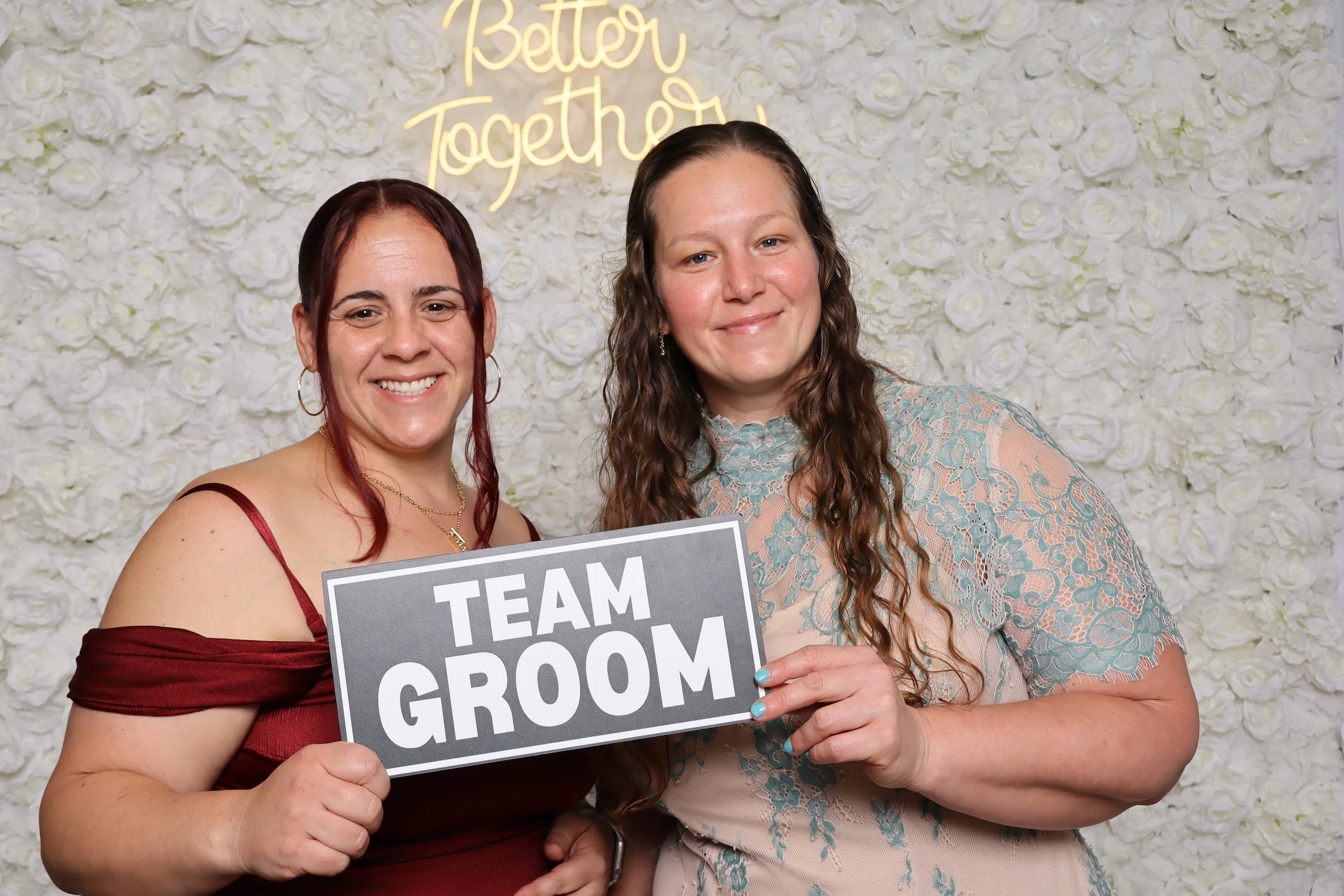 Two women smiling at a wedding photo booth, holding a sign that says 'TEAM GROOM'. The background is a white floral wall with a neon yellow sign that reads 'Better Together'.