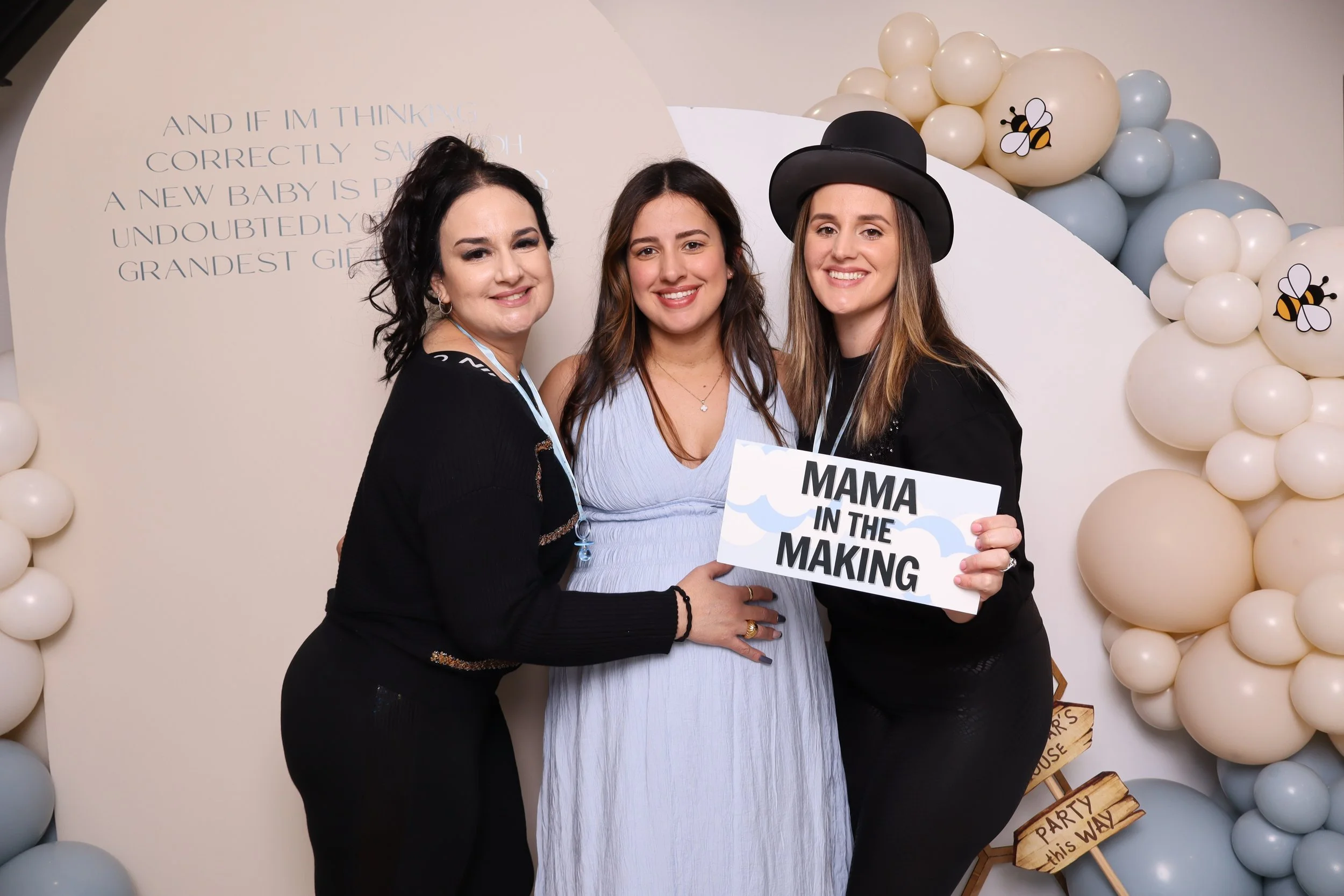 Three women posing together at a baby shower, with balloons and a sign that reads 'Mama in the Making'. The woman in the middle is pregnant and smiling, wearing a light blue dress. The woman on the right is holding a sign and wearing a black top hat,
