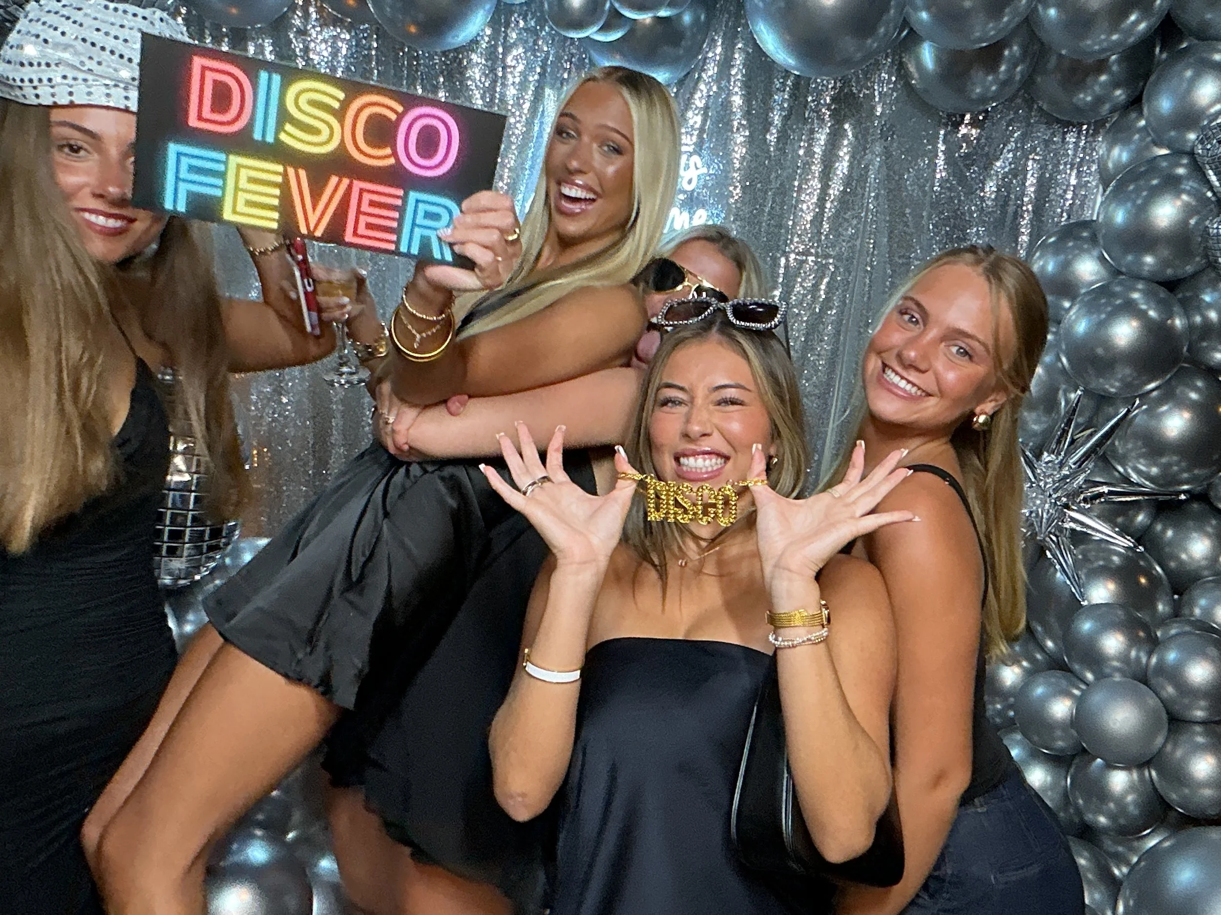 A group of five women celebrating at a party with a silver balloon backdrop, holding a colorful neon 'Disco Fever' sign, some wearing sunglasses and jewelry, and one wearing a 'Disco' necklace, smiling and making playful poses.