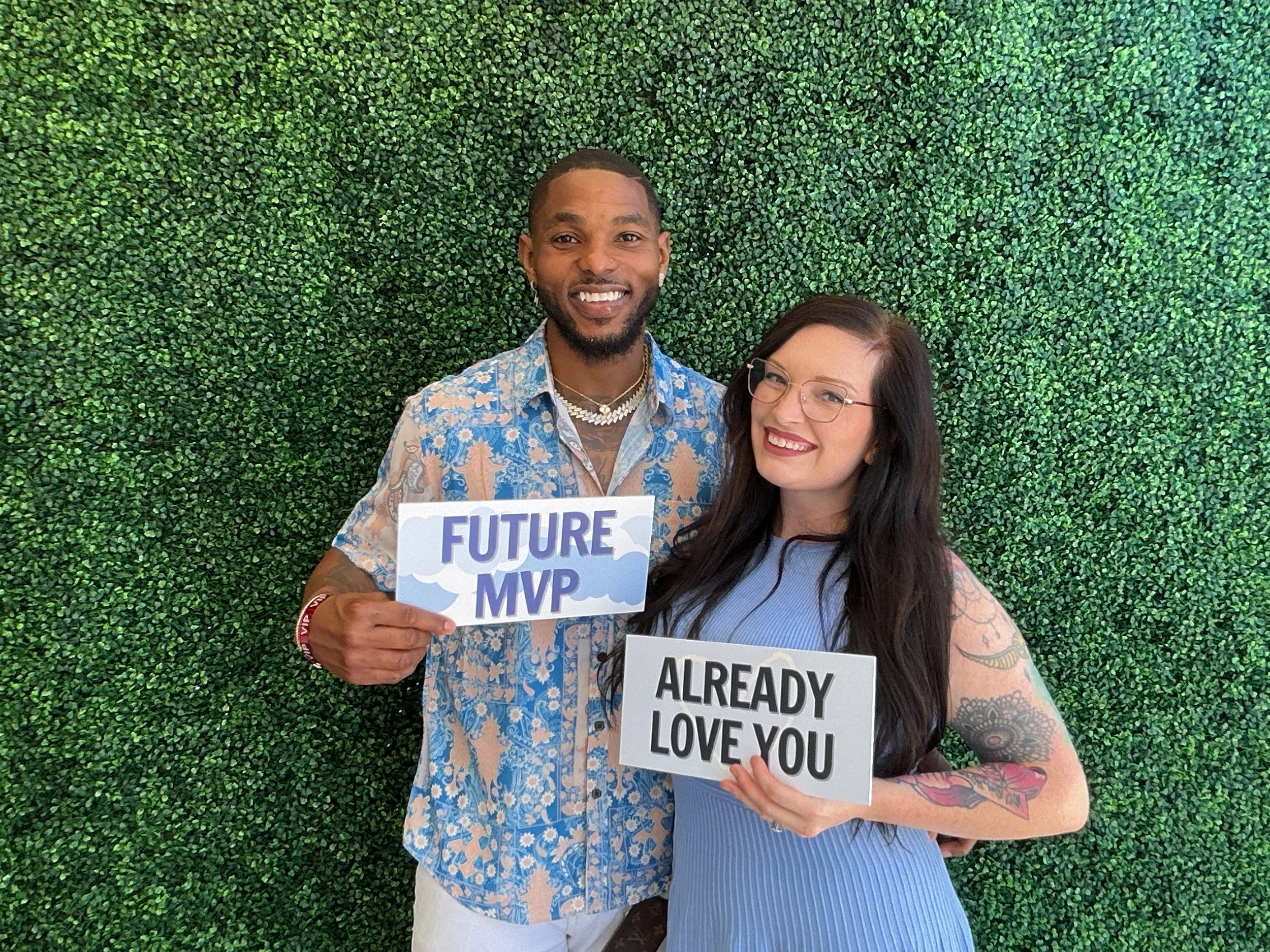 A smiling man and woman posing in front of a green leafy wall, holding signs that read "FUTURE MVP" and "ALREADY LOVE YOU," respectively.