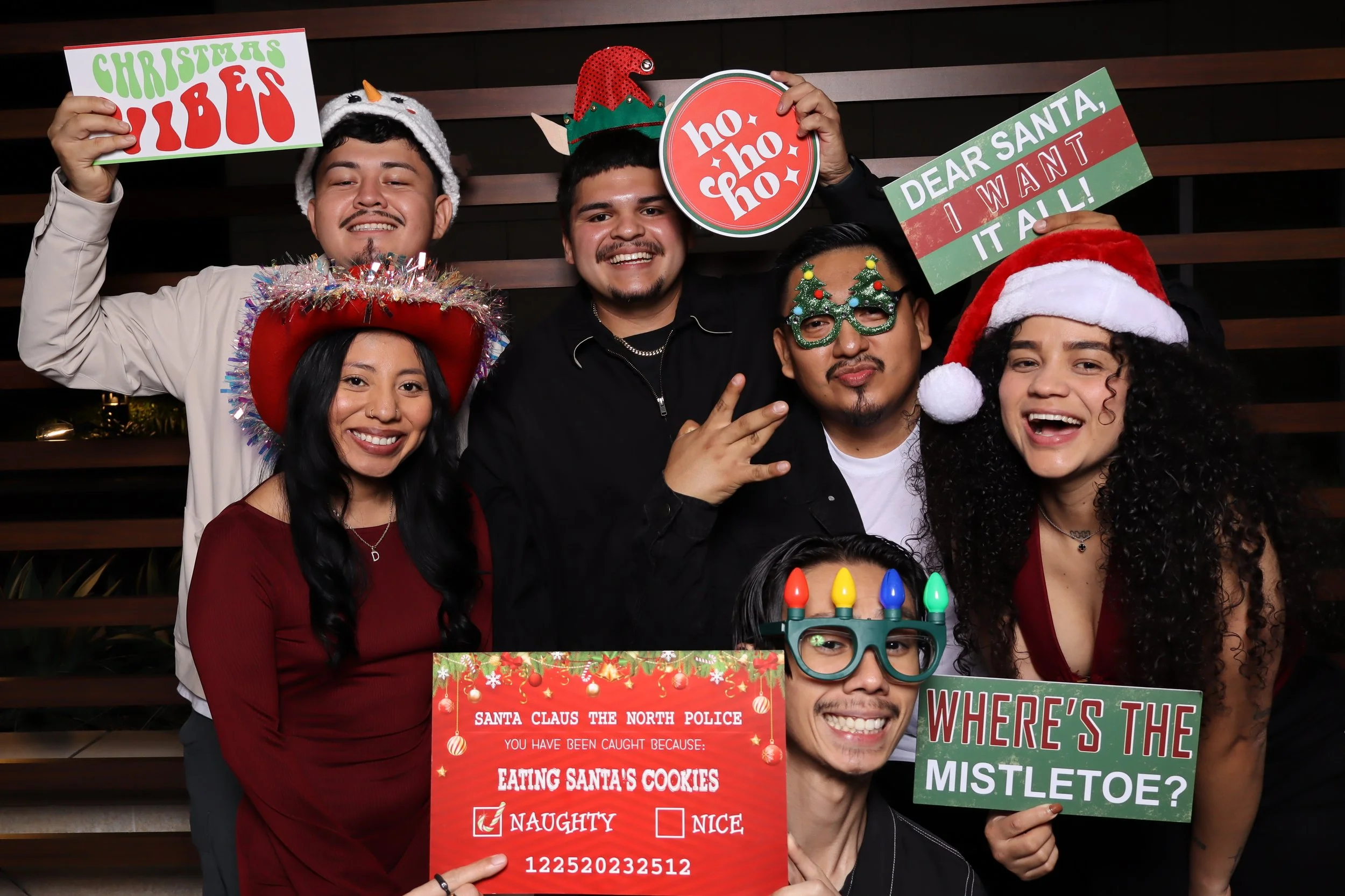 Group of six friends celebrating Christmas, wearing festive accessories like Santa hats, holiday glasses, and a Christmas hat. They are holding Christmas-themed signs and a humorous police certificate, with a wooden wall background.