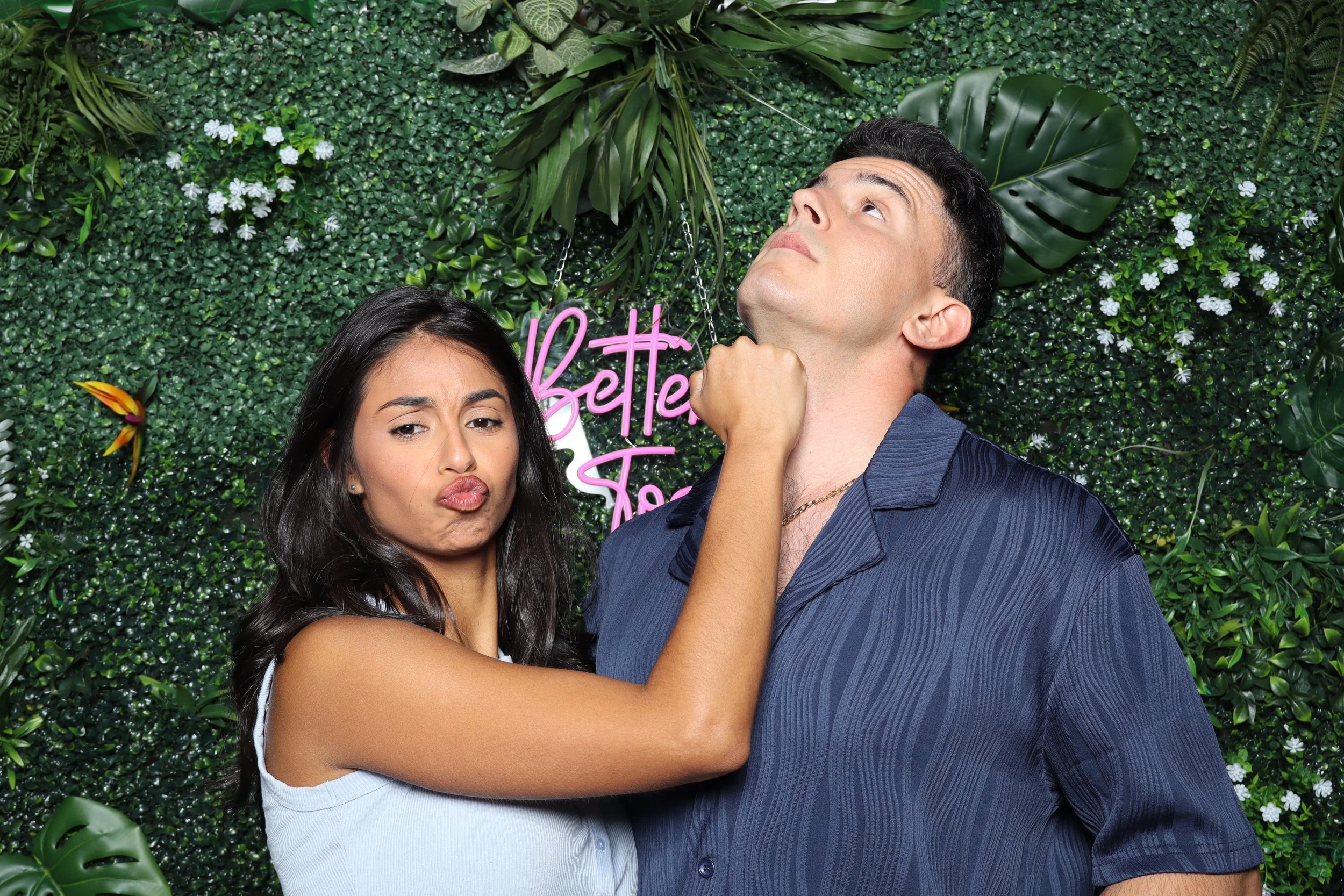 A woman with long dark hair making a playful face and a man with dark hair looking upward, with a green leafy foliage background and a pink neon sign that reads 'Bette To'.
