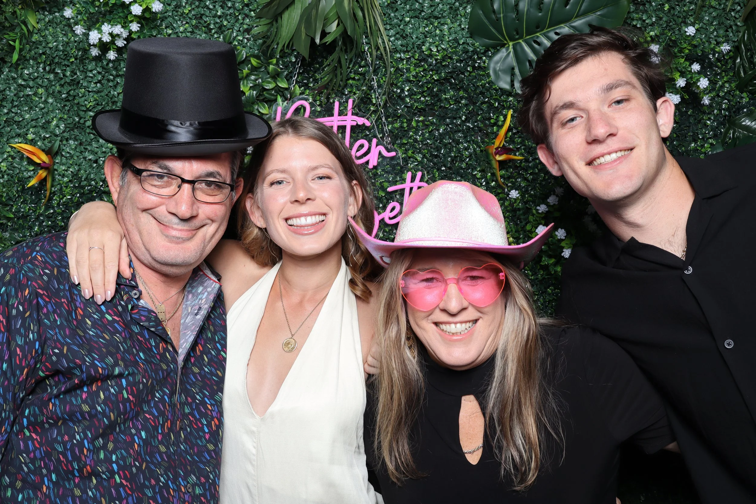 Group of four people smiling and embracing each other at a festive event, with a green leafy background and pink neon sign that says 'hotter than'.