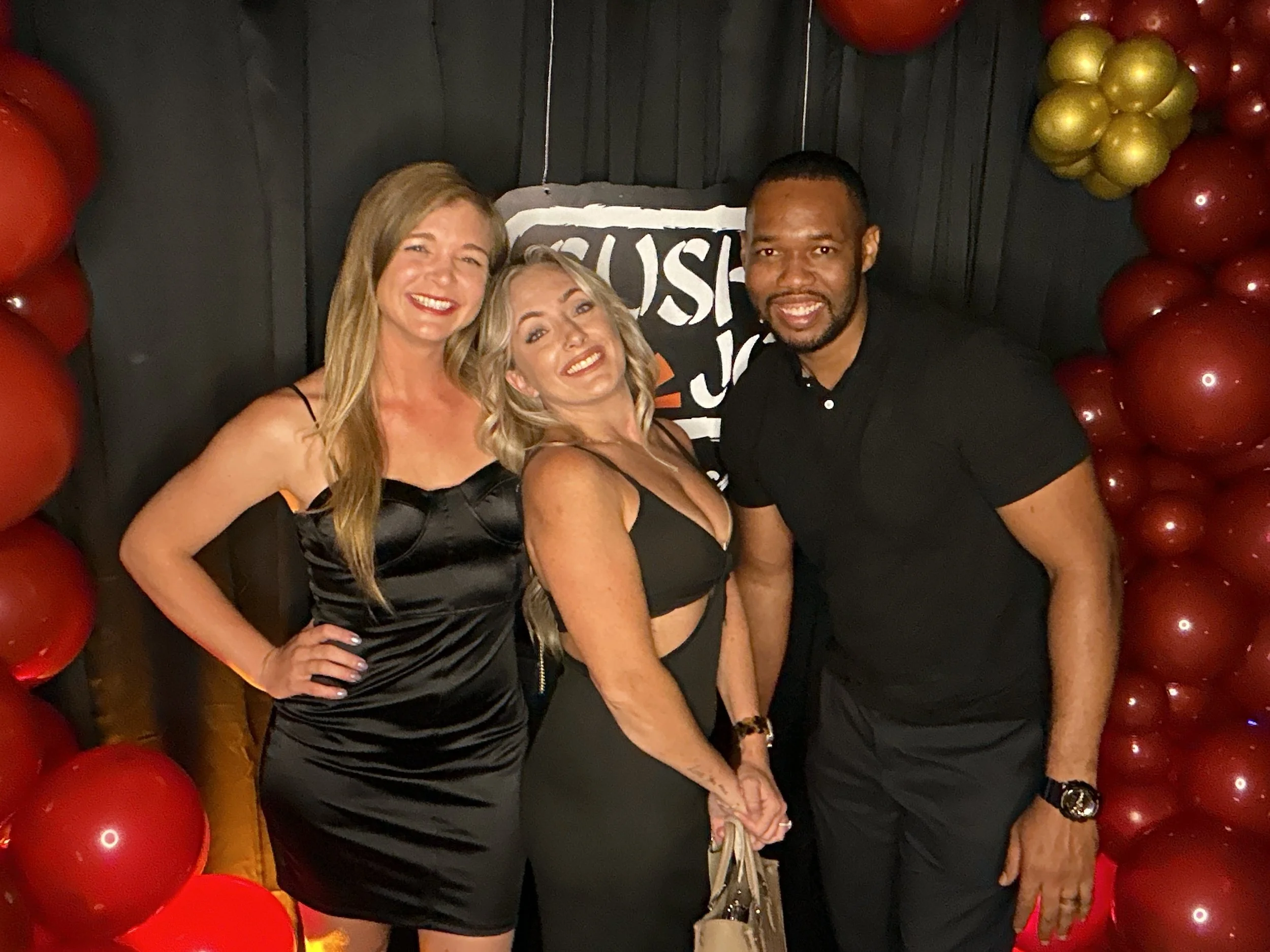 Three adults standing together in front of red and gold balloon decorations, smiling for the photo. The woman on the left is wearing a black satin dress, the woman in the middle is wearing a black dress with cutouts, and the man on the right is weari