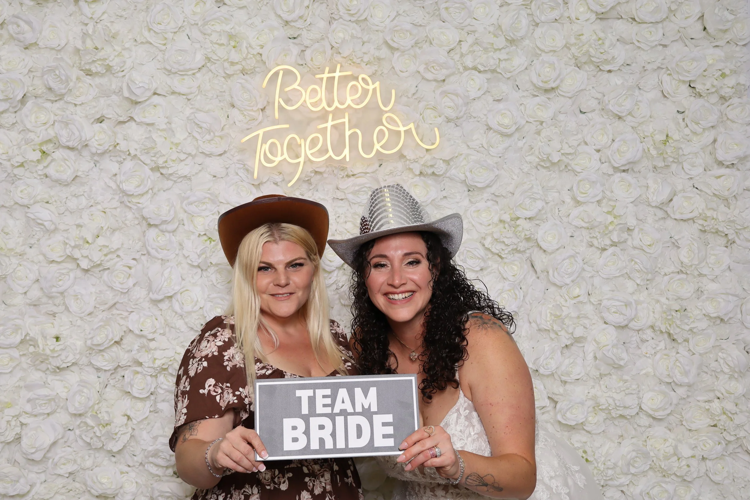 Two women in cowboy hats smiling at a wedding photo booth with a white rose wall background and a neon sign that says 'Better Together.' The woman on the right is holding a sign that says 'TEAM BRIDE'.