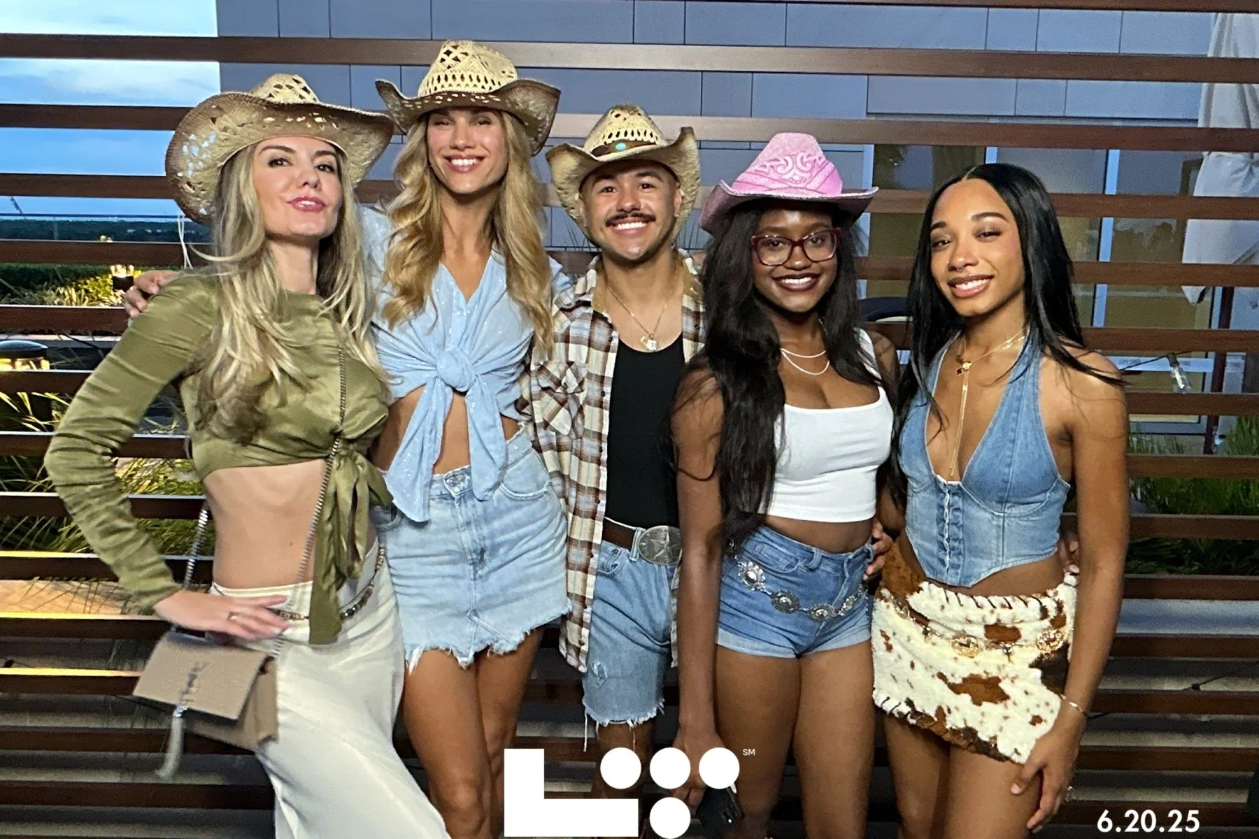 Group of five friends dressed in cowgirl and western-themed outfits, standing outdoors in front of a wooden fence, smiling at the camera during the evening.