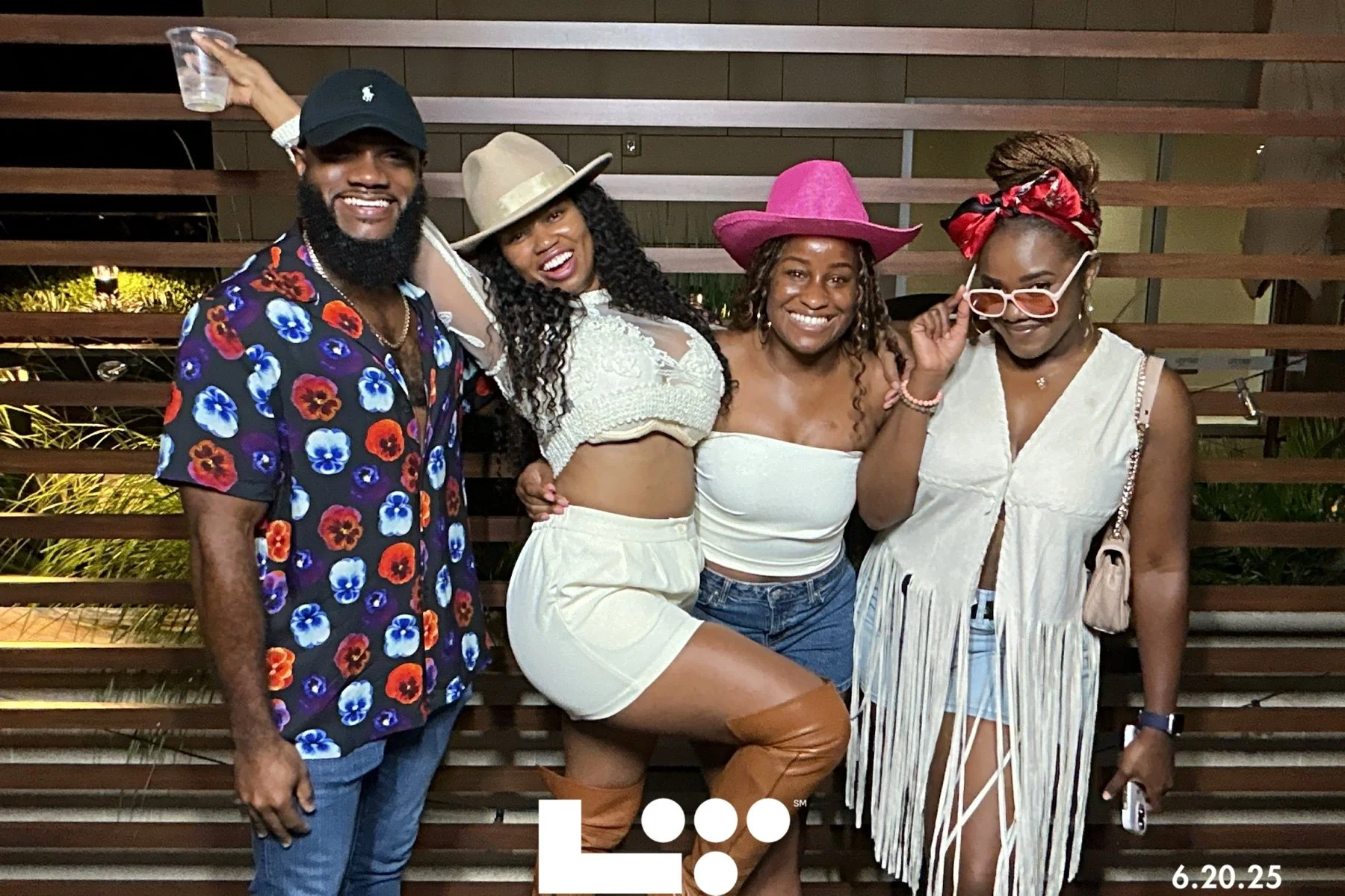 Group of four friends smiling and posing together at night outdoors. The man on the left is wearing a colorful floral shirt, a black cap, and has a beard. The second woman from the left is wearing a beige hat, white top, white shorts, and brown knee-