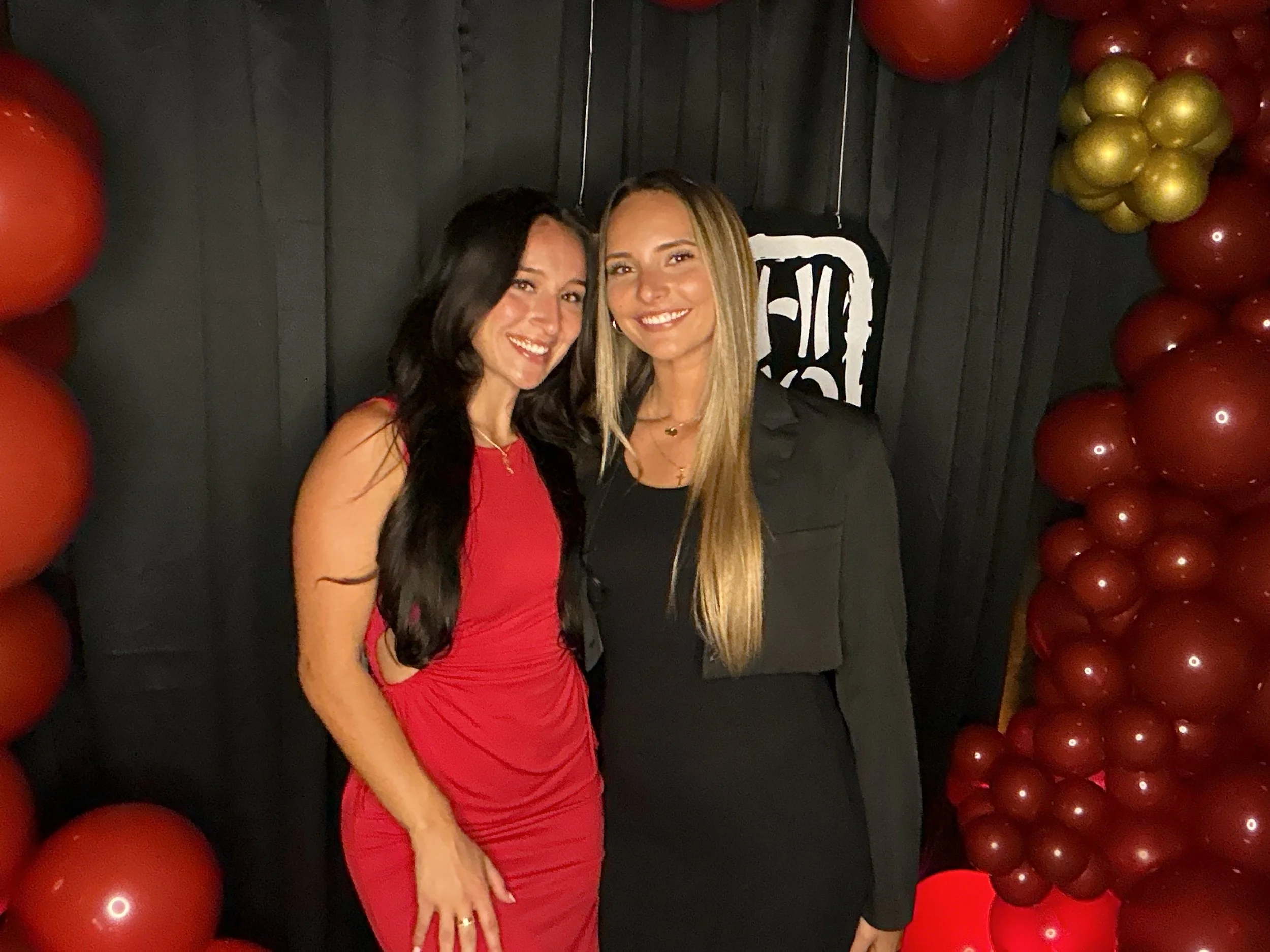 Two women standing close together at an event, smiling for the camera. One woman is wearing a red dress with long black hair, and the other is in a black outfit with long blonde hair. They are surrounded by red and gold balloons and a black backdrop 