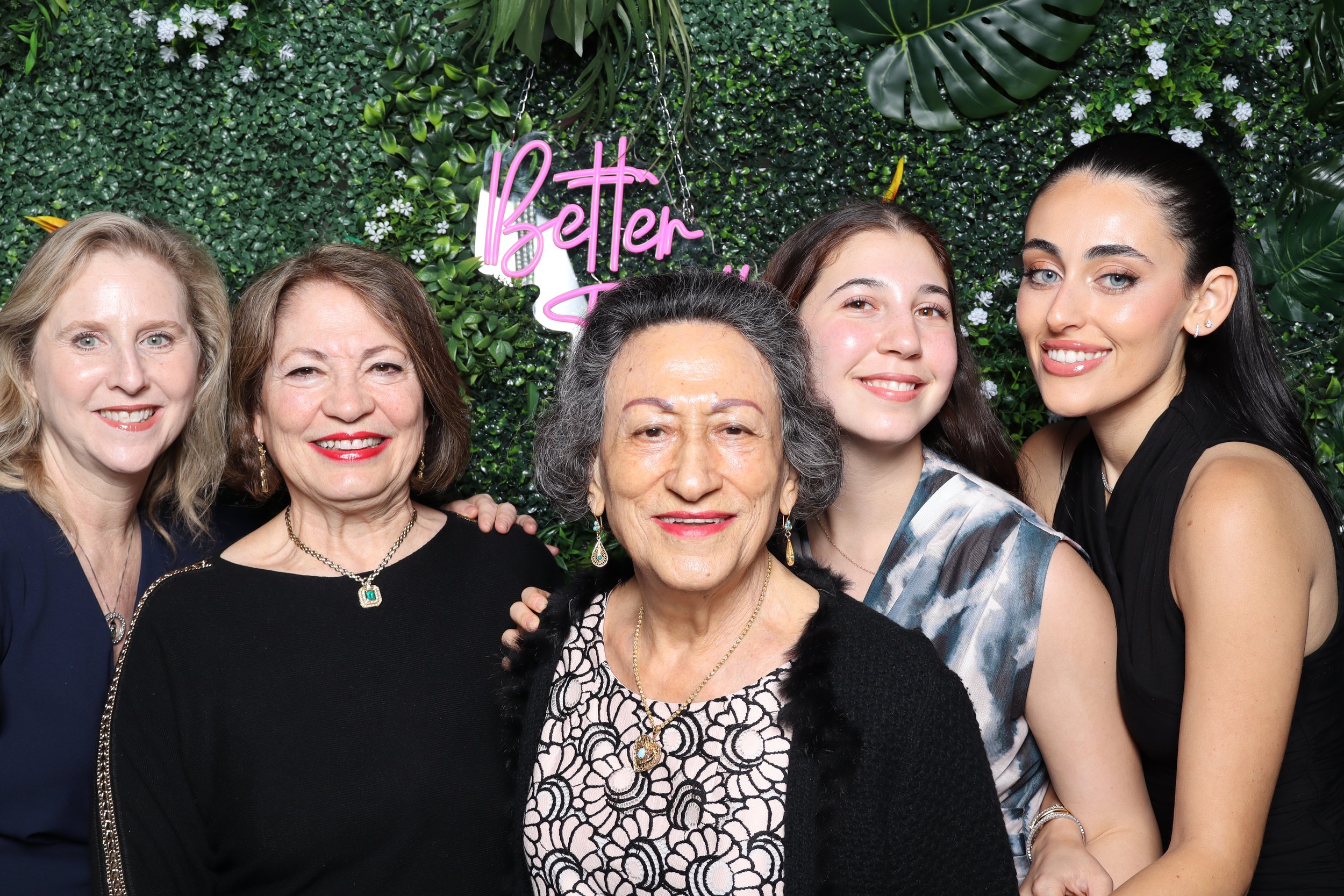 A multigenerational group of six women smiling at a celebration with a green plant wall background and a pink neon sign reading 'Better Together.'