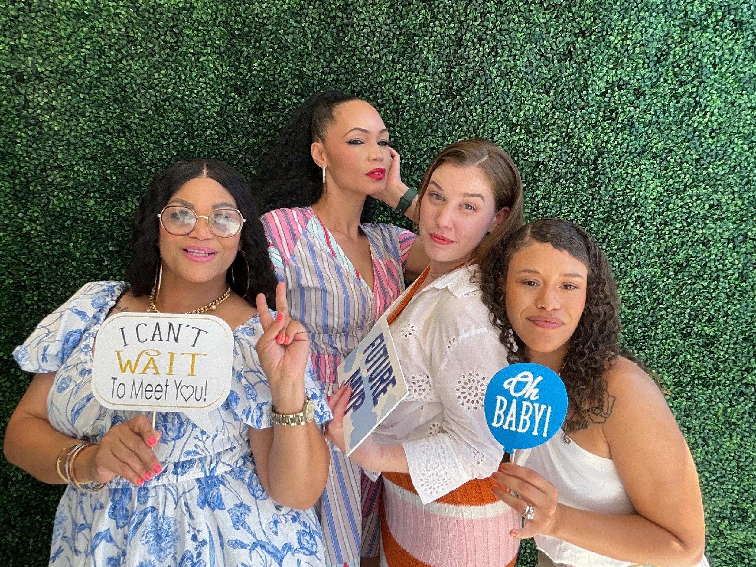 Four women posing in front of a green leafy background, holding signs with playful messages. The woman on the far left wears glasses and a blue and white floral dress, holding a sign that says 'I CAN'T WAIT To Meet You!'. The woman second from the le