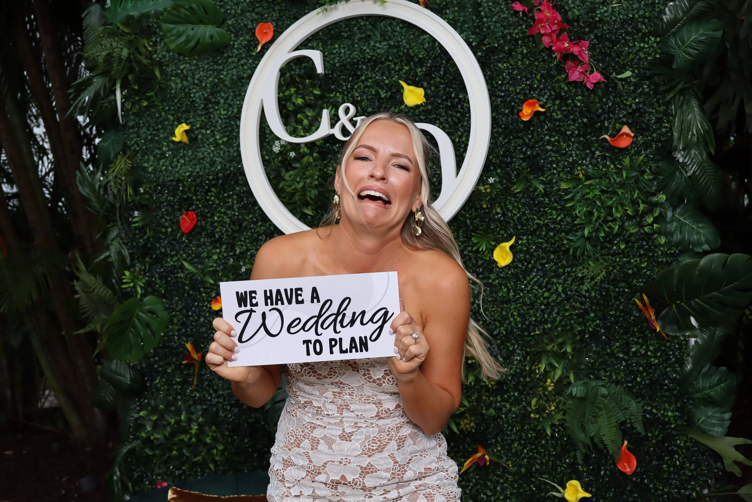 A woman in a strapless lace dress holding a sign that reads 'We have a wedding to plan', standing in front of a green foliage wall with flowers and a circular wedding sign in the background.